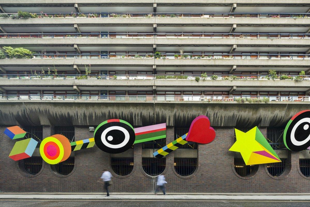A large brick structure and a pavement below a block of high-rise flats. Several colourful objects are imposed on the brick structure.