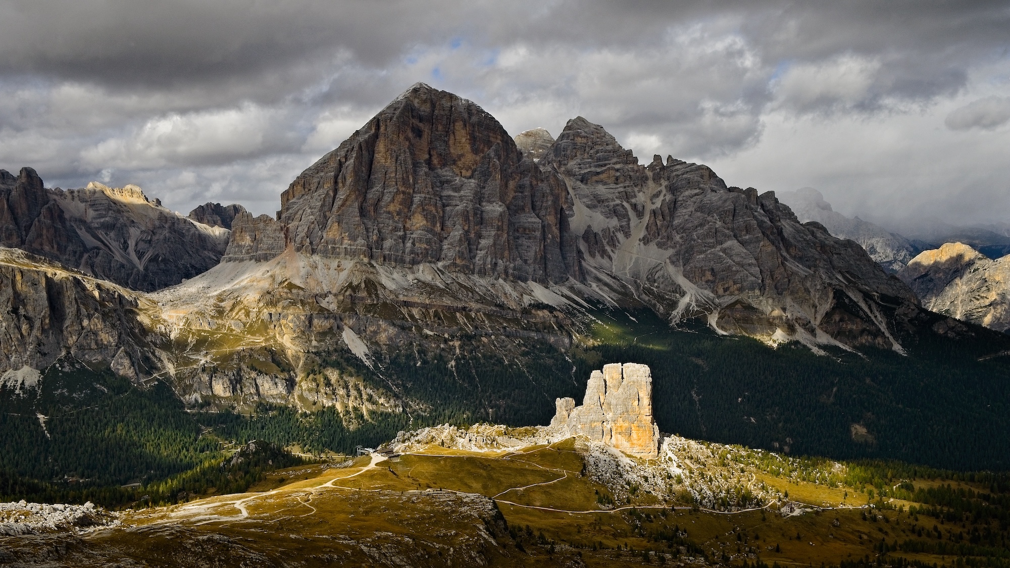 A panoramic view of the Dolomites showcasing tall, rugged mountains under a partly cloudy sky. Sunlight illuminates a rock formation in the foreground, while the surrounding peaks and valleys feature a mix of greenery and rocky terrain.