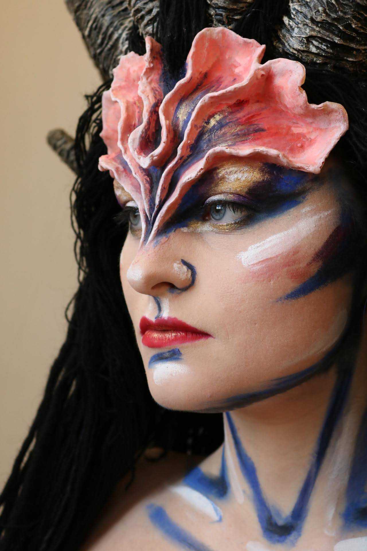Close-up headshot of a model facing to the left wearing blue and white war paint-style make-up and prosthetics on their forehead.