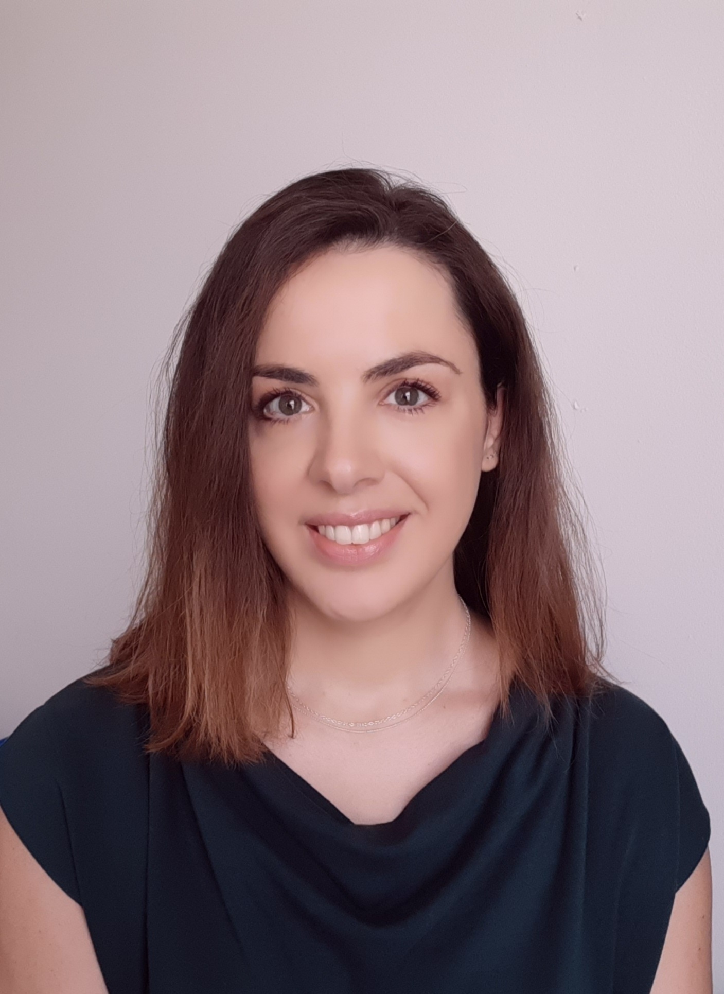 Woman with shoulder-length brown hair wearing dark blue top, smiling at camera against pale purple background.