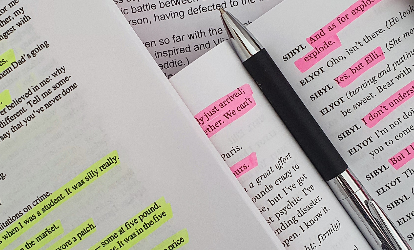Close-ups of scripts lying on top of one another. A pen rests in the centre fold of one. Lines have been highlighted in yellow and pink.