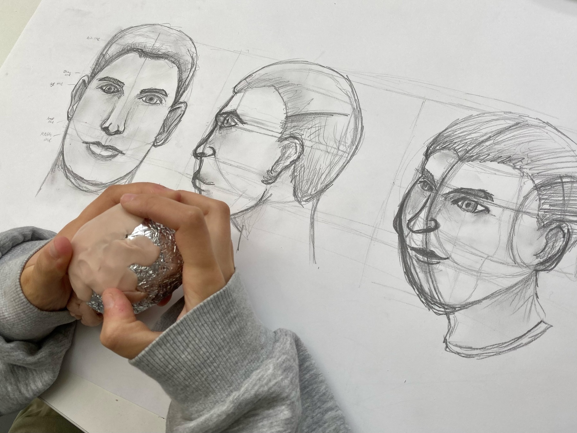 Hand with rings holding pencil over white paper with three pencil portrait sketches of men's faces in various angles.