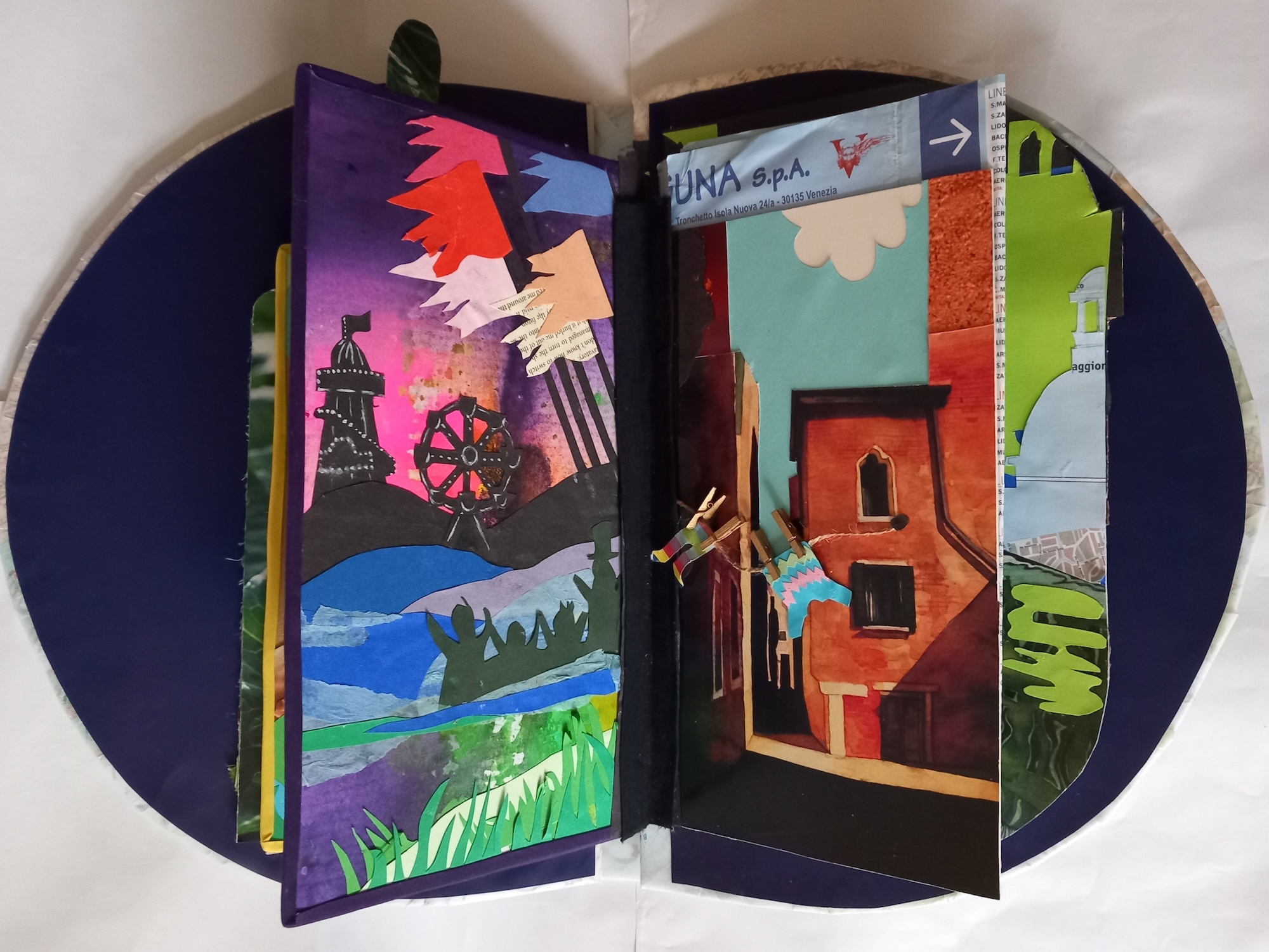 Open accordion-style book with colorful illustrated panels showing landscapes, buildings, and abstract shapes in blues, greens, reds, and purples on dark circular surface