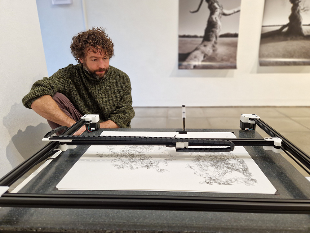Man with curly hair in green jumper operating large black printing press machine in gallery space with artwork on white walls.