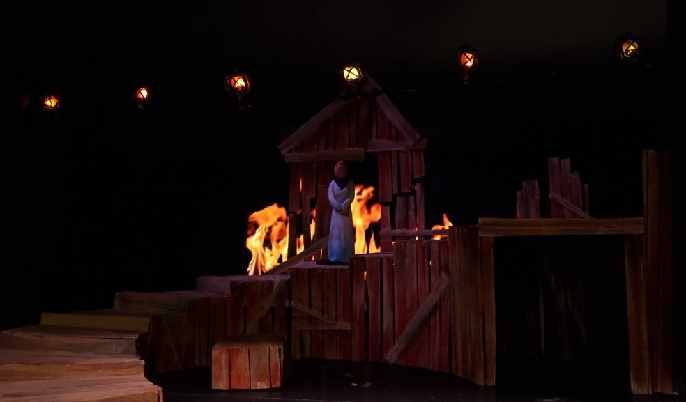 A model of a set design with wooden structures and fire in the background.