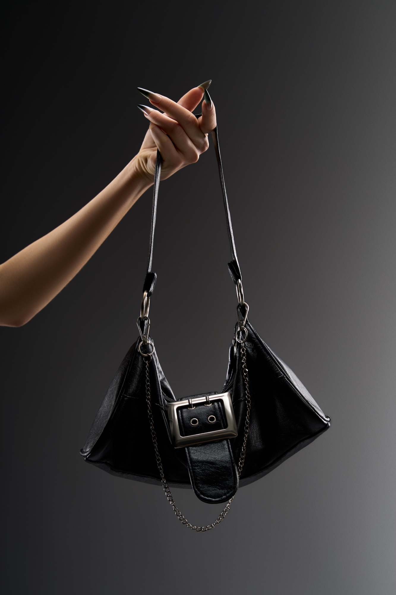 Hand holding black leather handbag with silver buckle and chain details against grey gradient background.