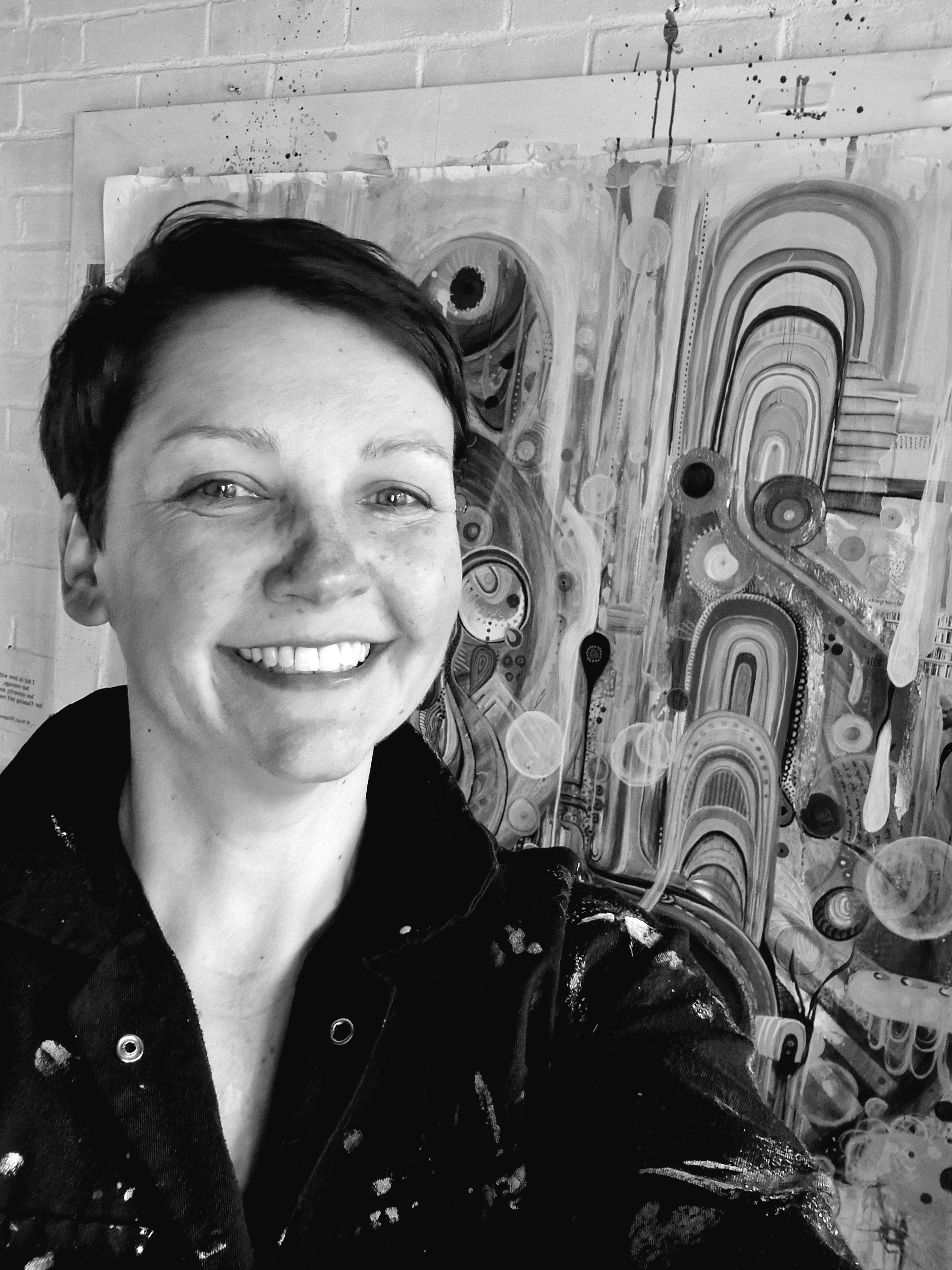 Woman with short dark hair smiling in black and white image, standing before abstract artwork with curved lines and circular shapes.