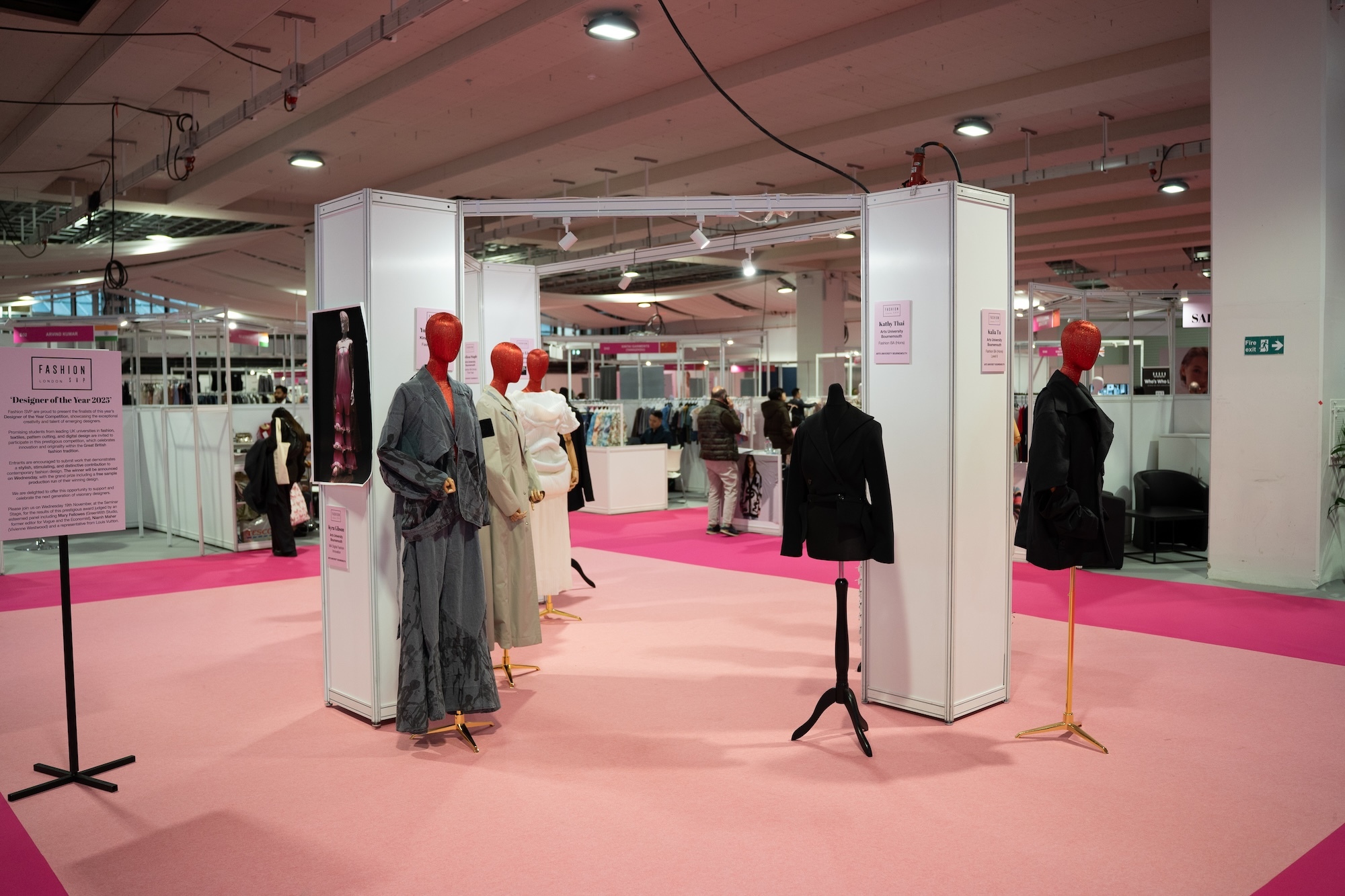 Exhibition hall with white entrance archway, pink carpet, and mannequins displaying various clothing including grey and black garments.