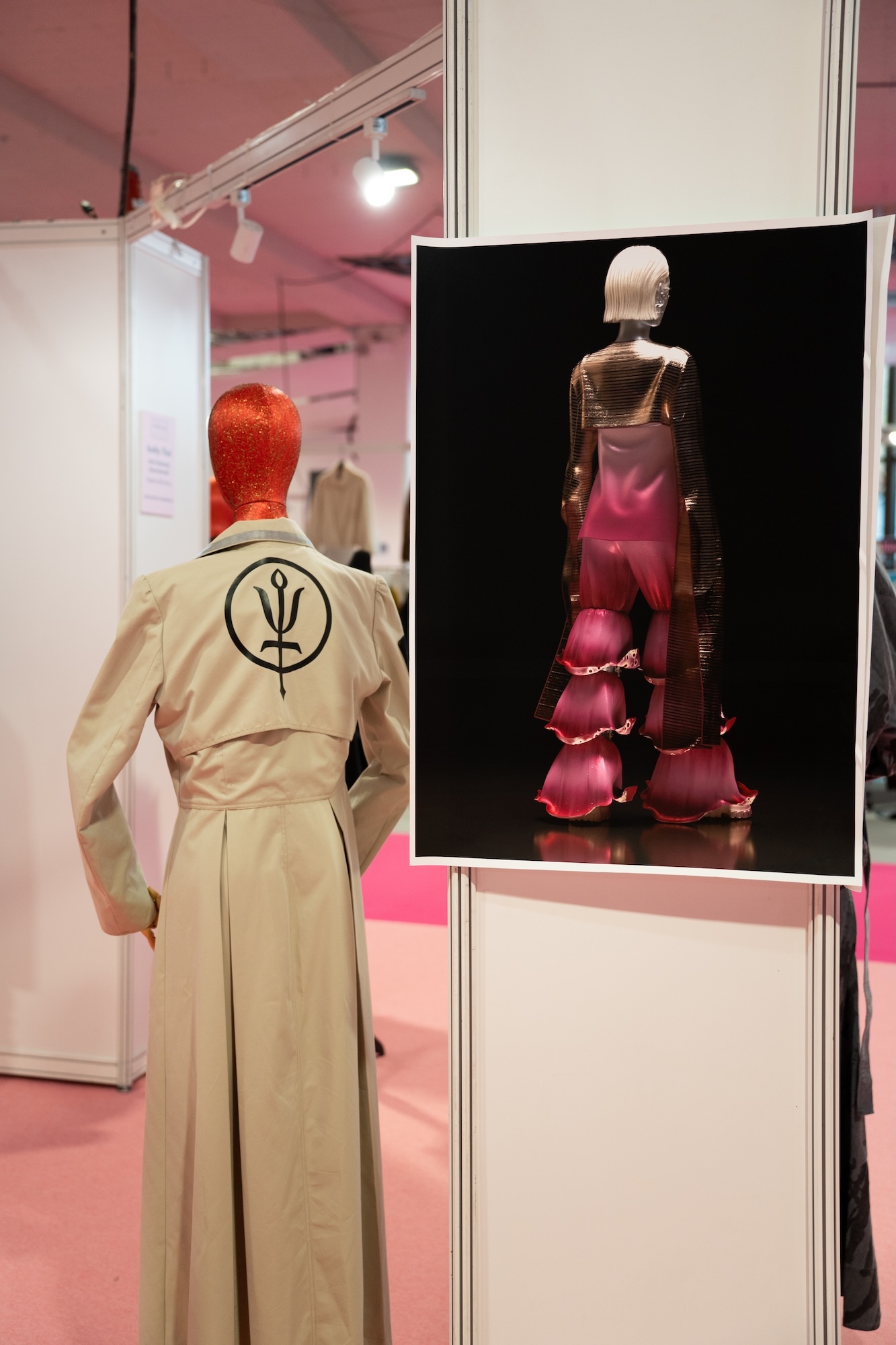 Museum display with beige trench coat on mannequin and framed image of pink ruffled dress against black background. Pink ceiling visible.