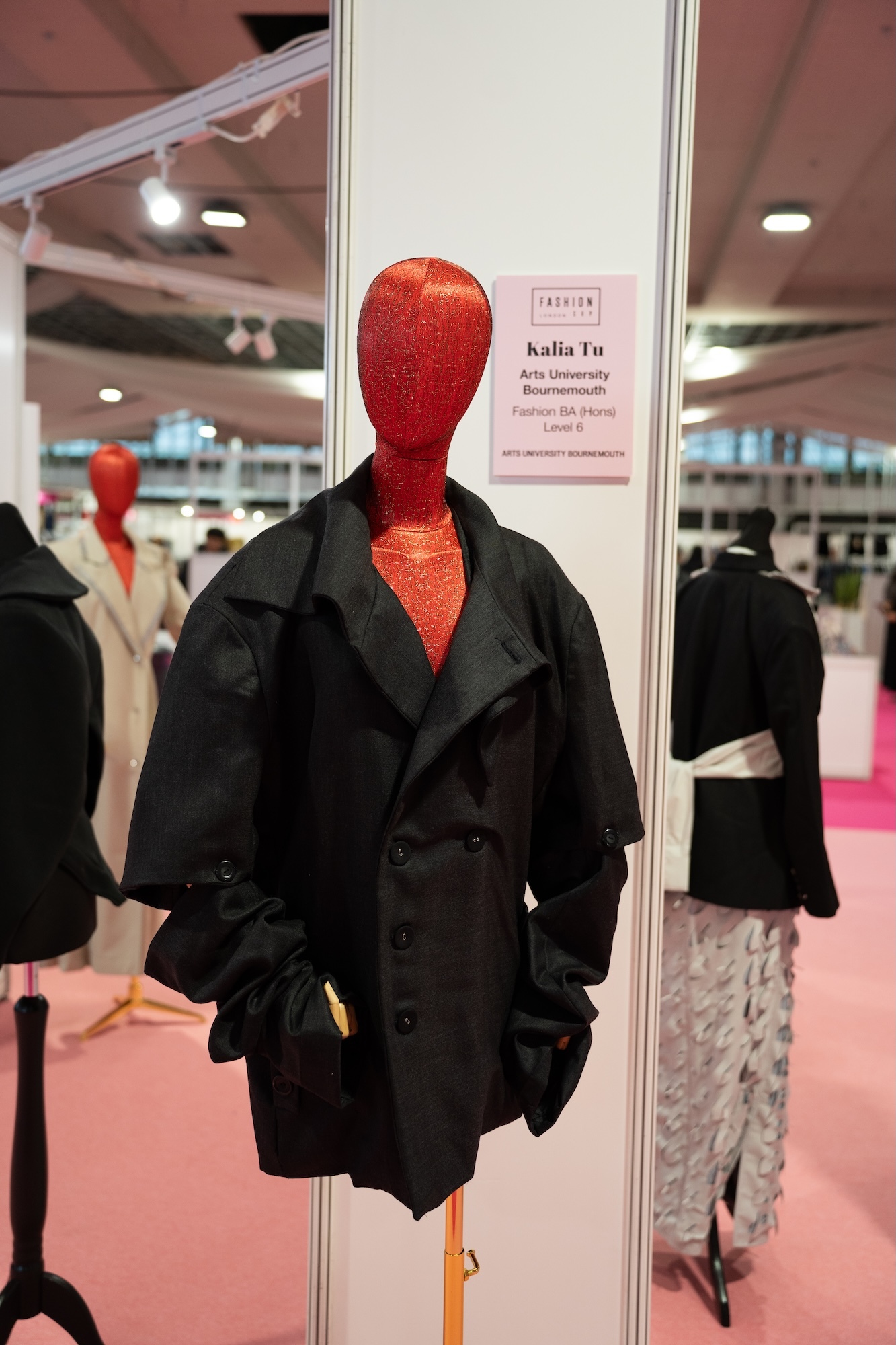 Red mannequin head wearing black double-breasted coat in glass display case at fashion exhibition with pink carpeted floor.