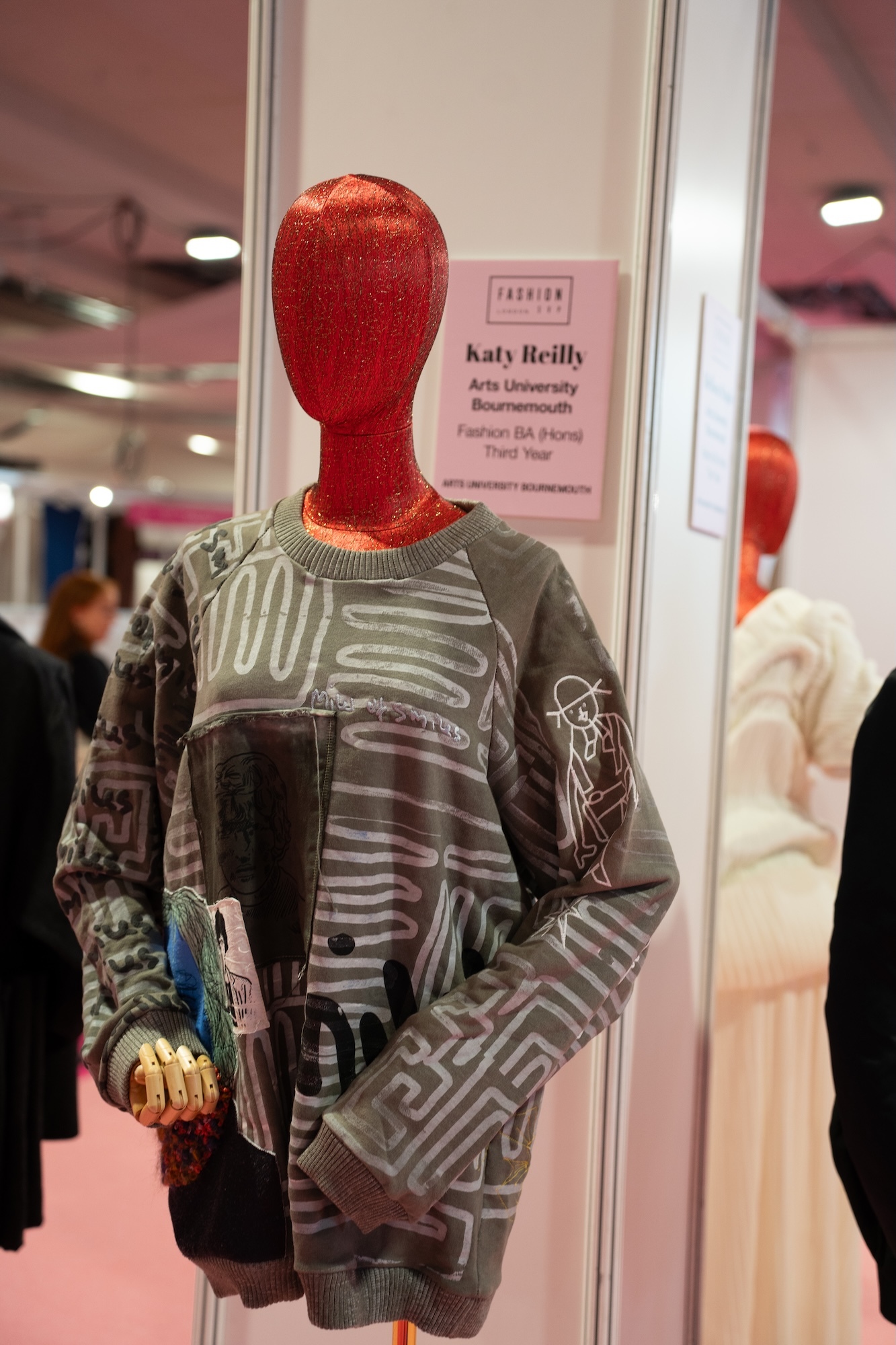 Mannequin with red metallic head wearing grey patterned sweatshirt in glass display case with name placard reading "Katy Reilly"
