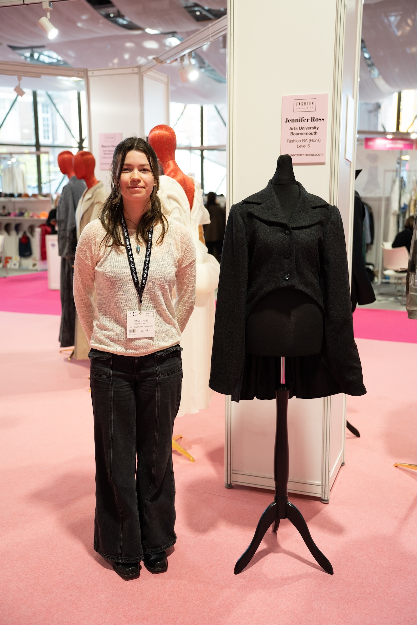Woman with lanyard stands beside black coat on mannequin at trade show with pink carpet and white exhibition stands.