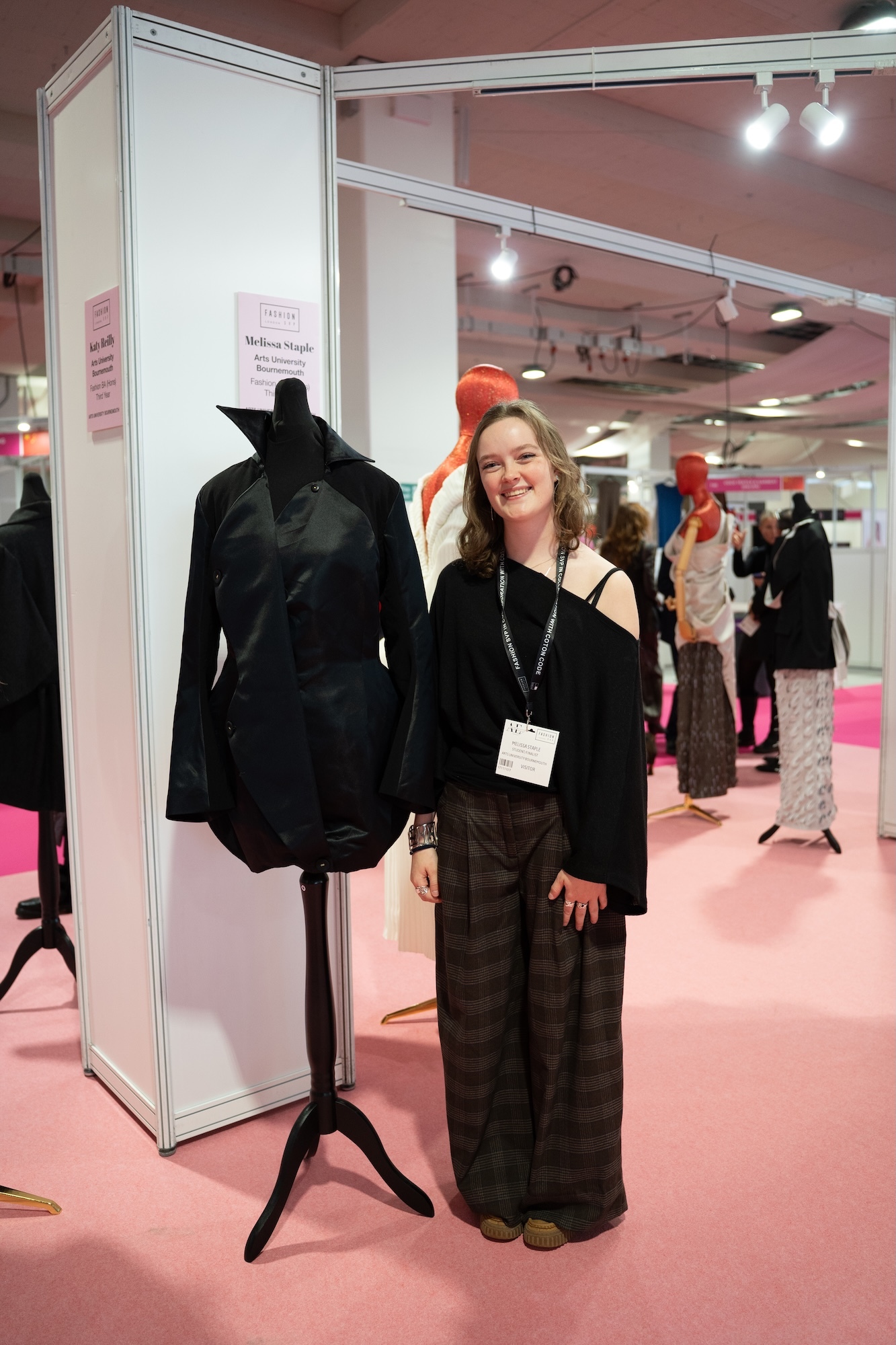 Woman with badge standing next to black jacket on mannequin at trade show booth with white display panels and pink flooring.