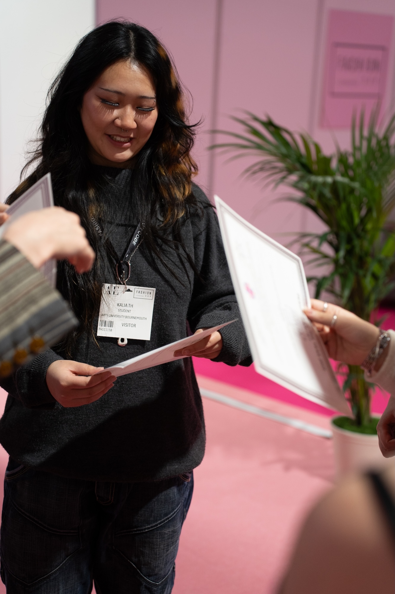 Woman with long dark hair wearing black jumper and name badge holds papers whilst speaking to someone off-camera in pink room.