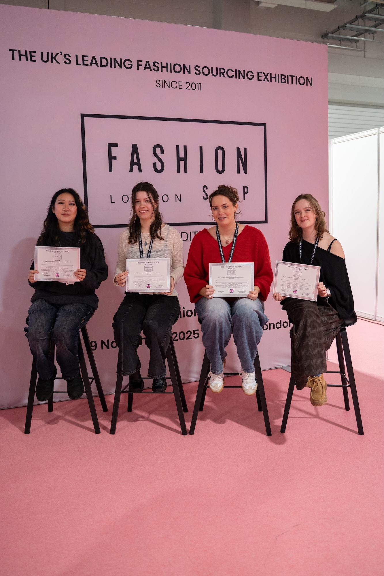 Four BA (Hons) Fashion students seated on black stools holding certificates at Fashion Sourcing London exhibition, pink backdrop with black text and logo.