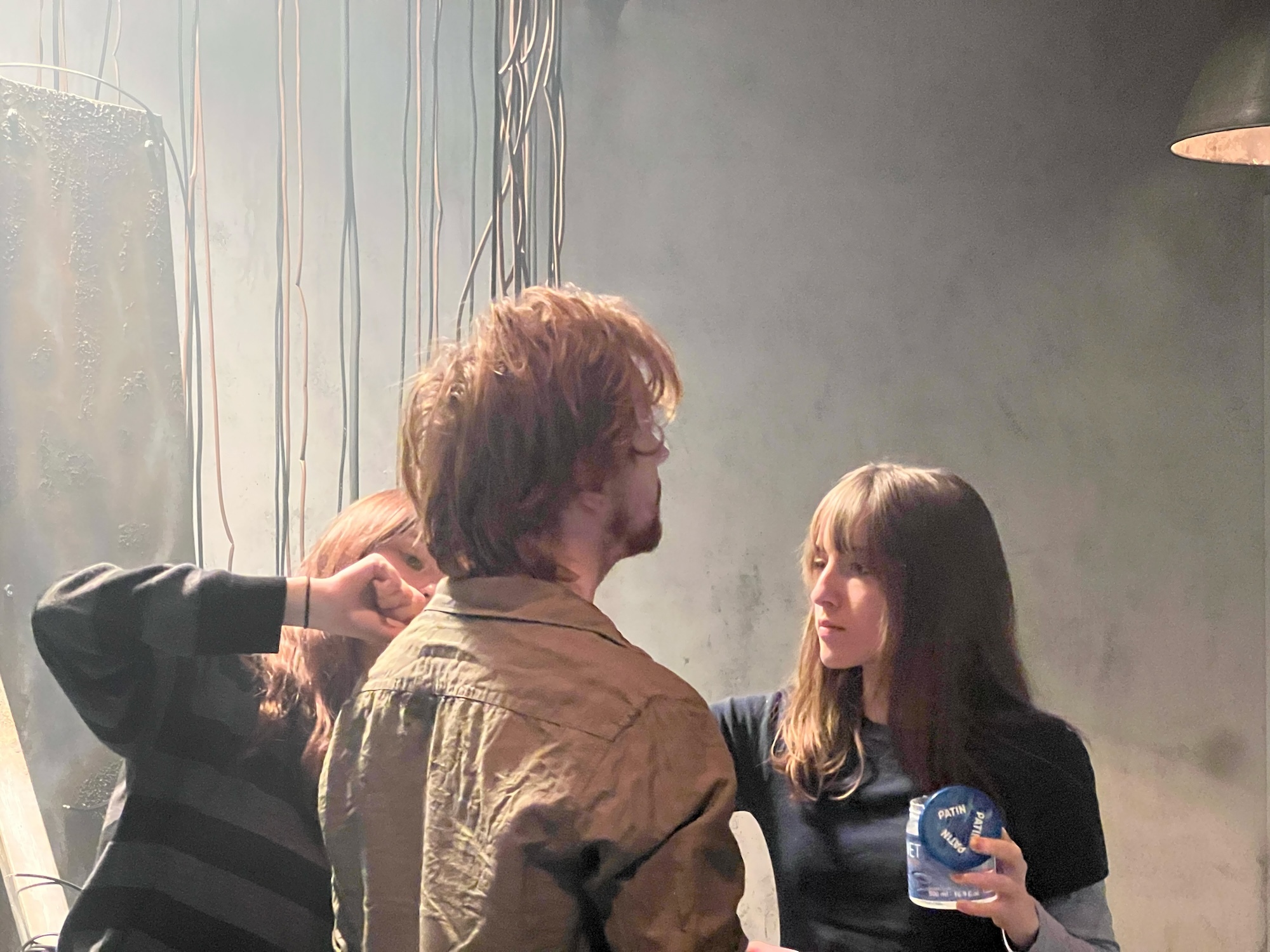 Man in brown jacket and woman in dark clothing standing near window with natural light streaming in, creating atmospheric haze.