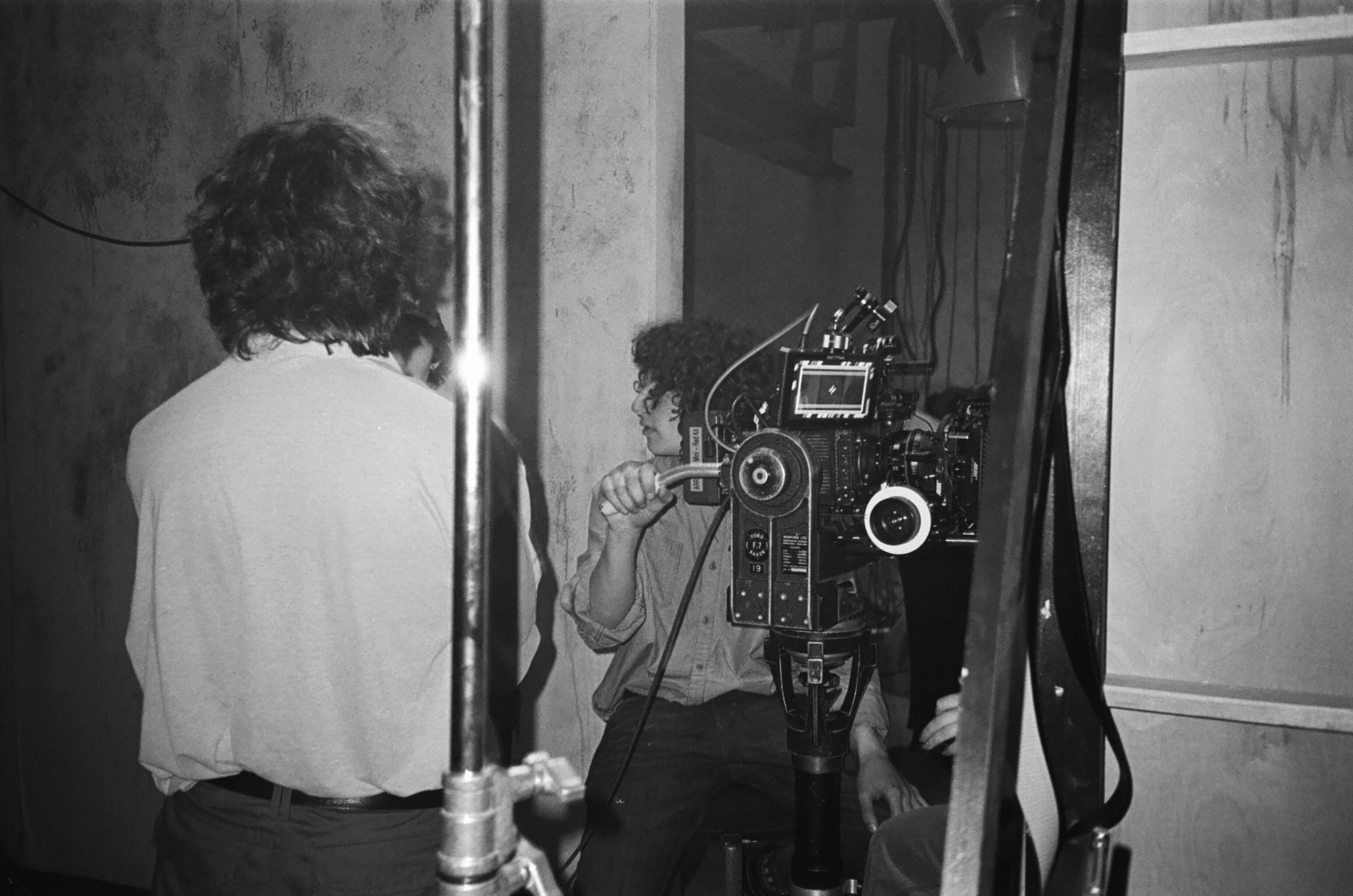 Black and white image of two people operating a film camera in an indoor setting with windows and architectural details visible.