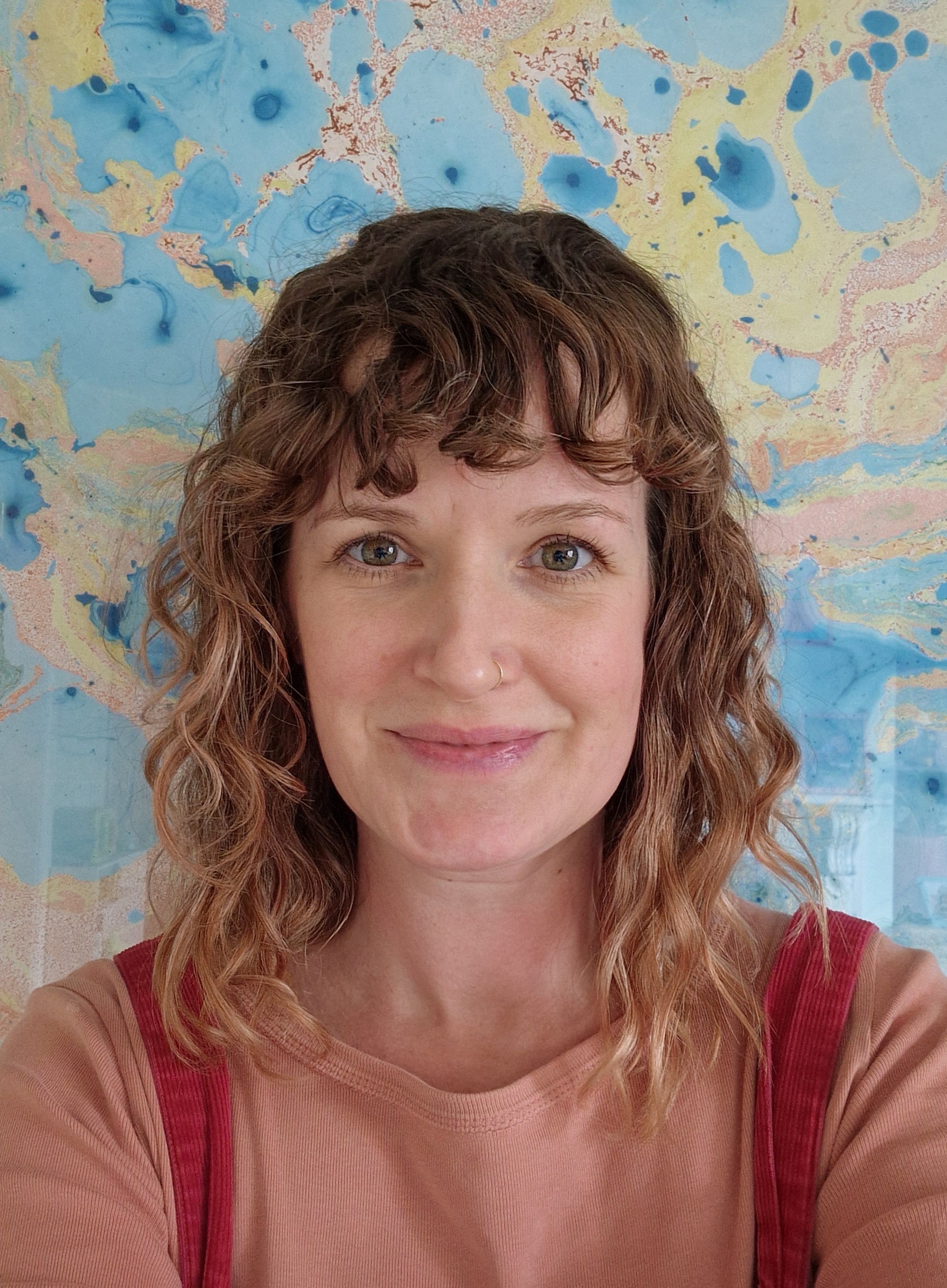 Woman with curly brown hair and fringe wearing red top, against blue and beige paint-splattered wall background.