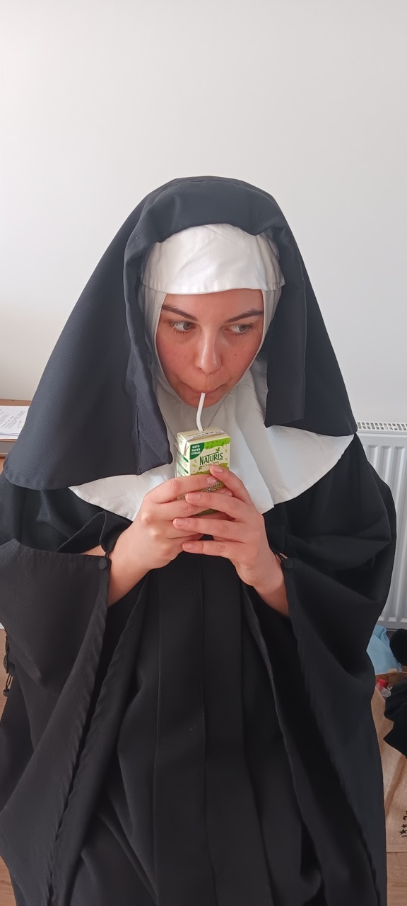 Person in black and white nun's habit drinking from small green and white carton through straw, sitting indoors.
