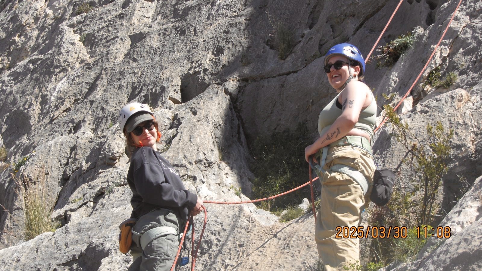 Two rock climbers with helmets and ropes on grey limestone cliff face, one wearing dark clothing, other in beige trousers.