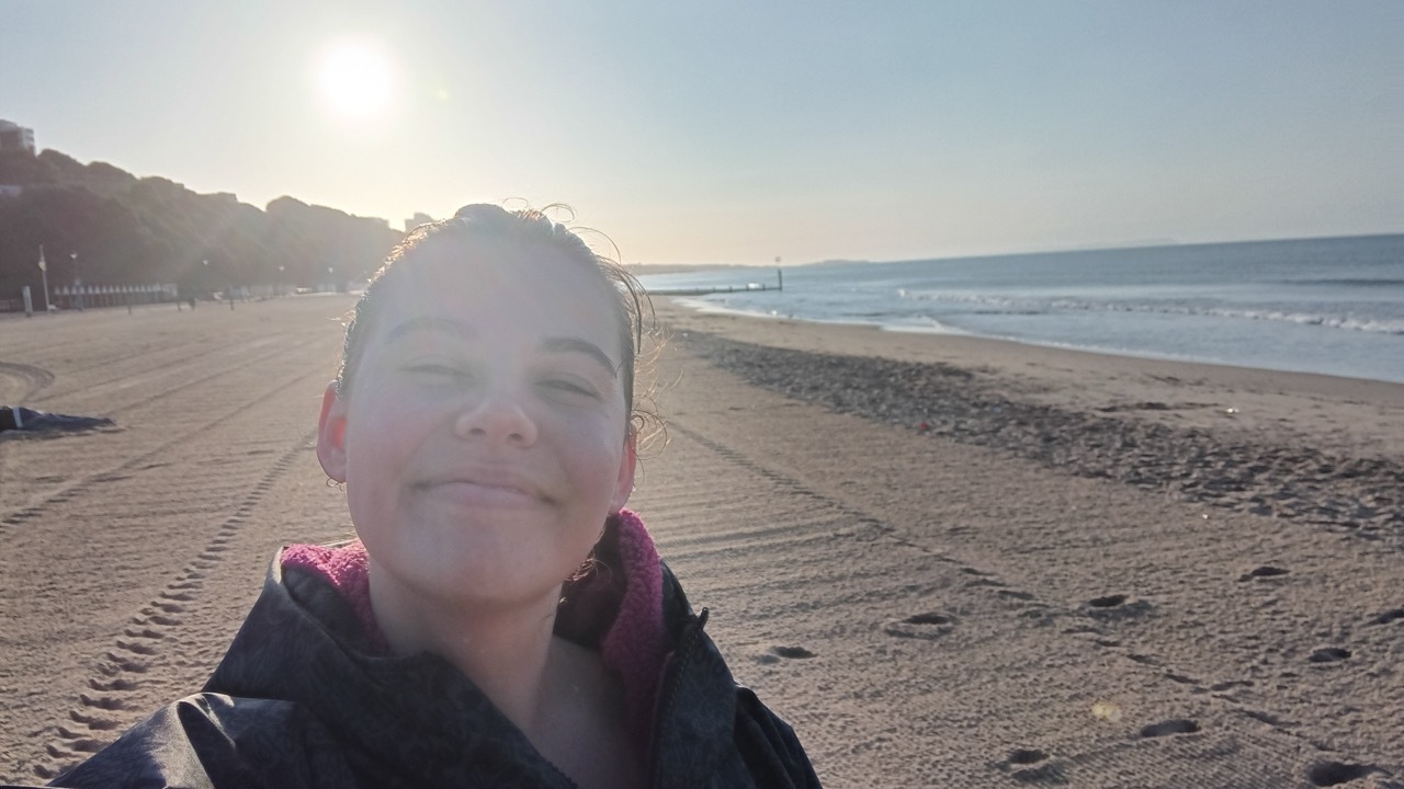 Person wearing dark jacket smiling at camera on sandy beach with bright sun creating lens flare, ocean and coastline in background.