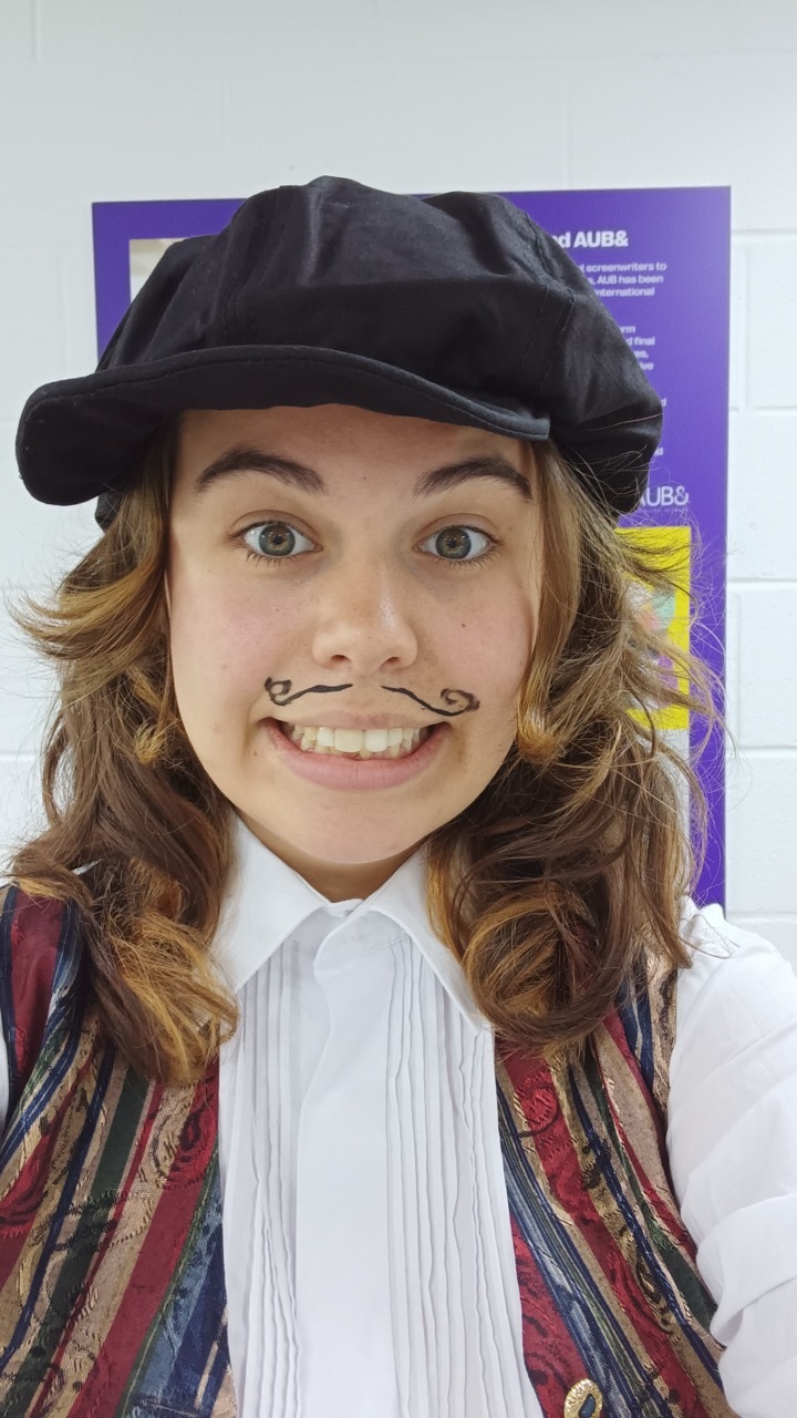 Woman with blonde hair wearing black cap, white shirt, and striped waistcoat with drawn-on moustache, smiling at camera.