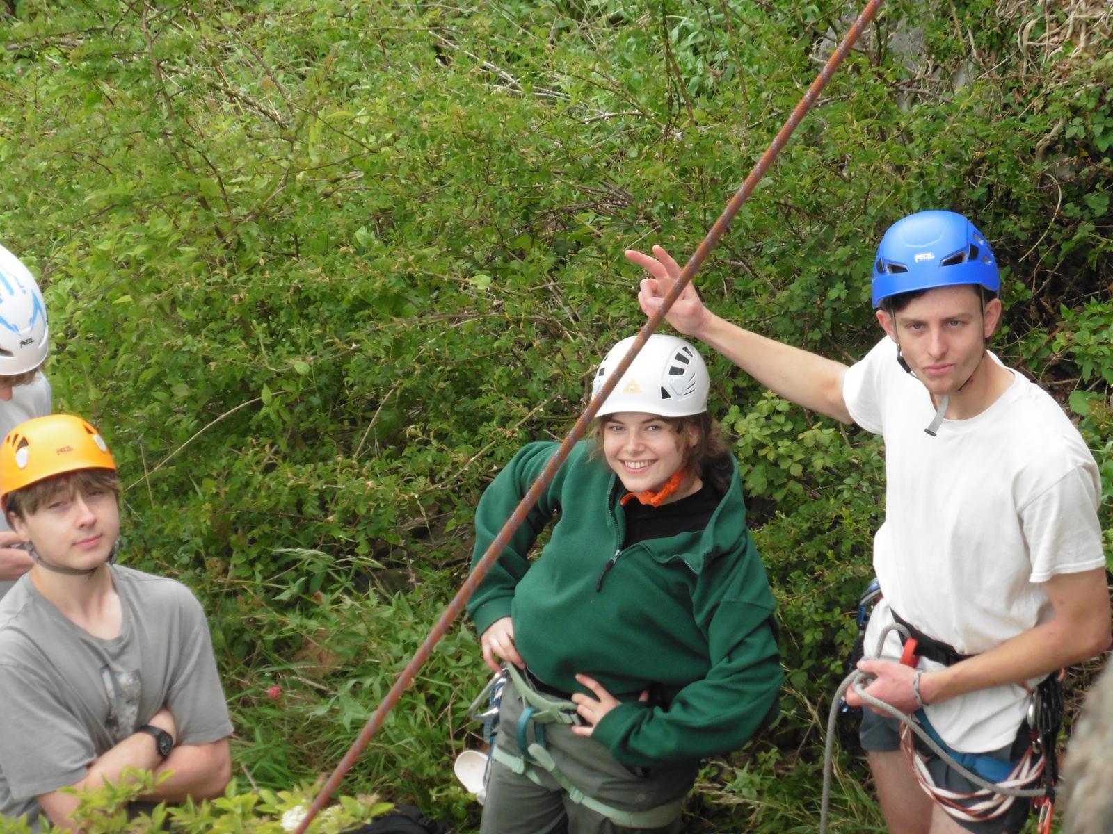 Three people in safety helmets and harnesses during outdoor climbing activity, surrounded by green foliage and trees.