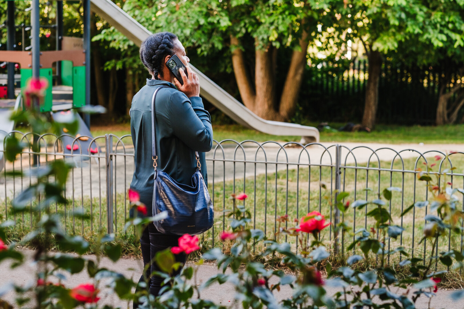 A woman with a shoulder bag speaks on a mobile phone beside a flower bed in a park, with a playground slide and trees in the background.
