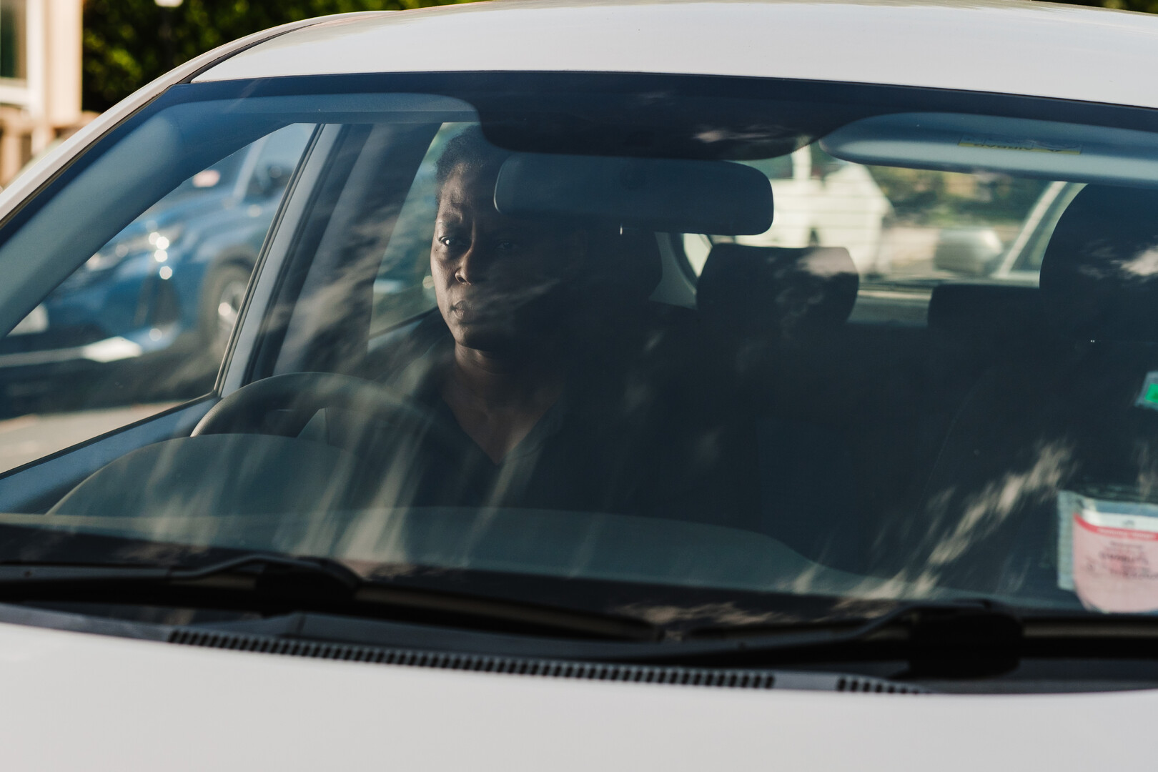 A man sits in the driver's seat of a white car, seen through the front windscreen with reflections on the glass.