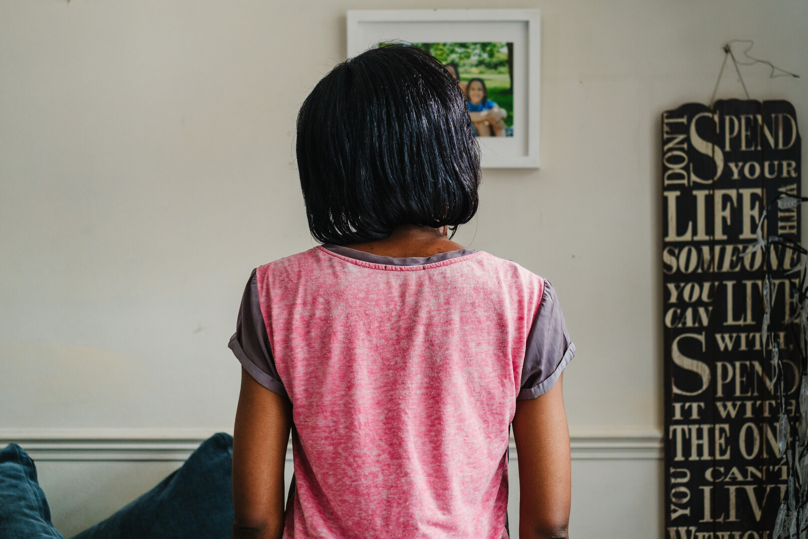 A woman with short black hair stands with her back to the camera, wearing a pink T-shirt in a room with a framed photo on the wall and a black typographic hanging sign.