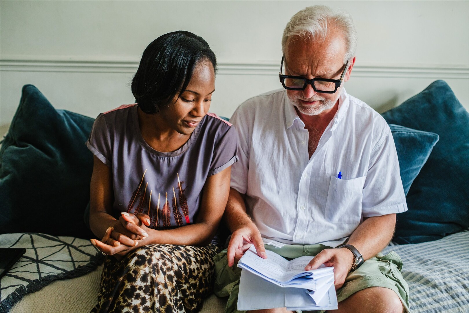 A Black woman and an older white man sit side by side on a sofa, looking at an open book, with dark blue cushions behind them.
