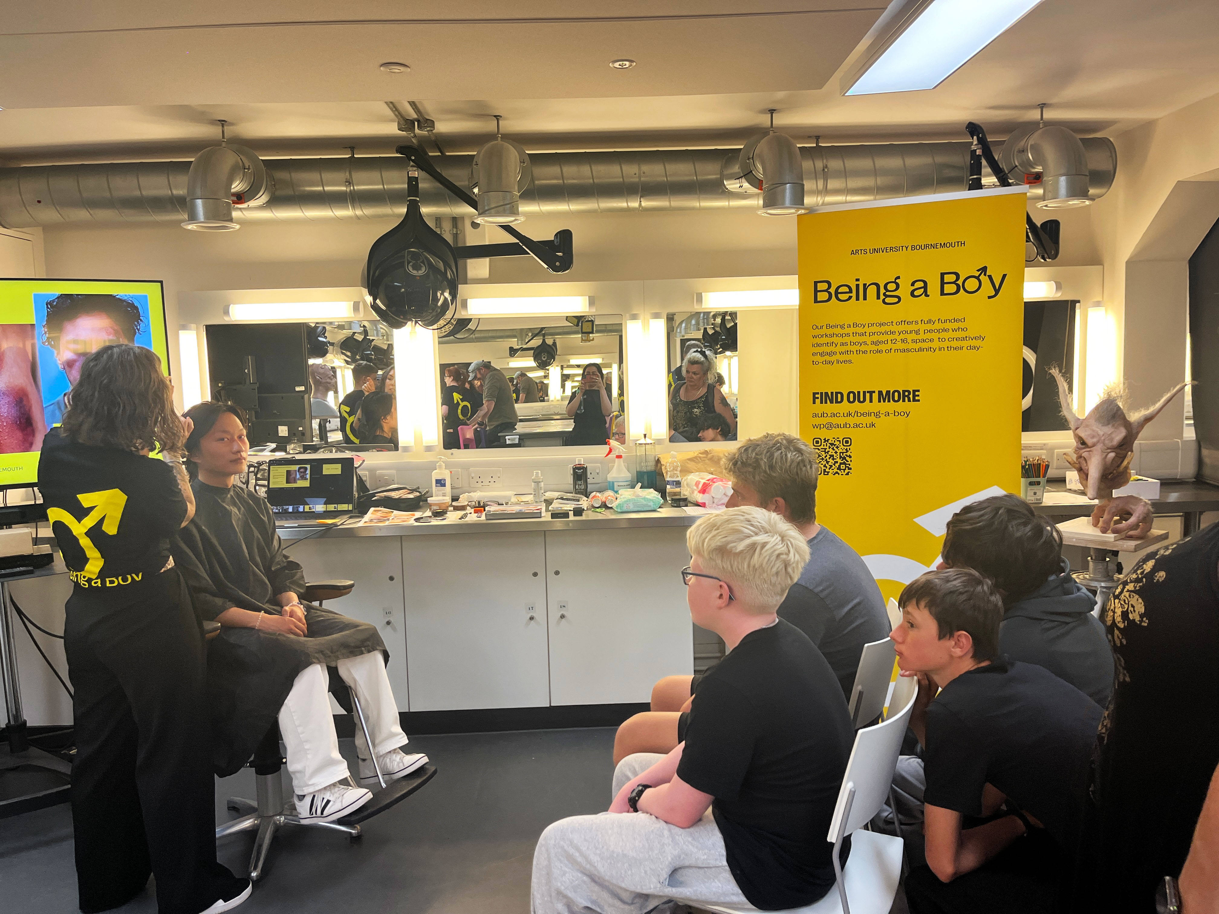 Make-up studio interior with customers in chairs, staff cutting hair, yellow "Being a Boy" promotional banner hanging from ceiling.