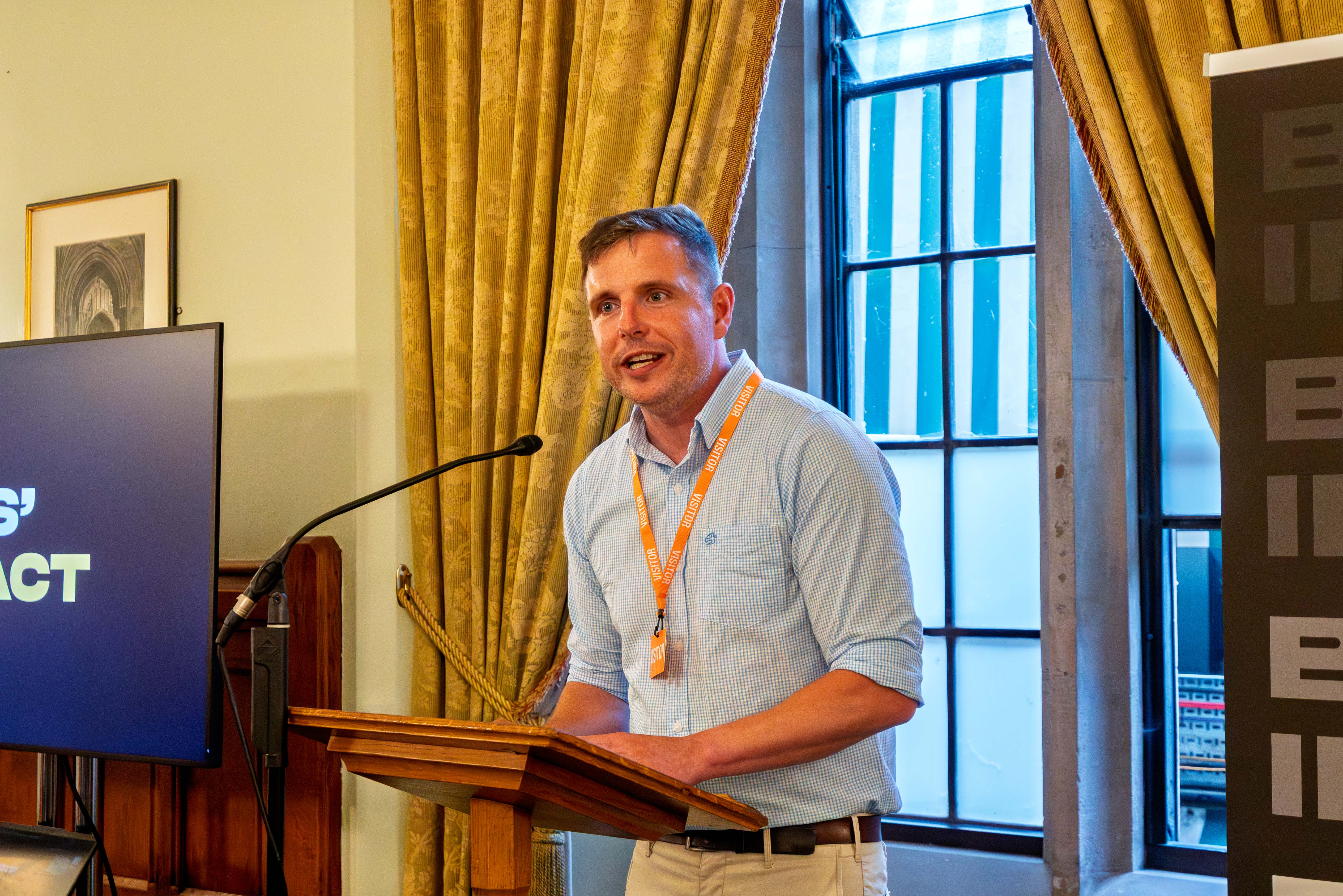 Dr Alex Blower at House of Commons speaking at a podium, screen, curtains and window behind.