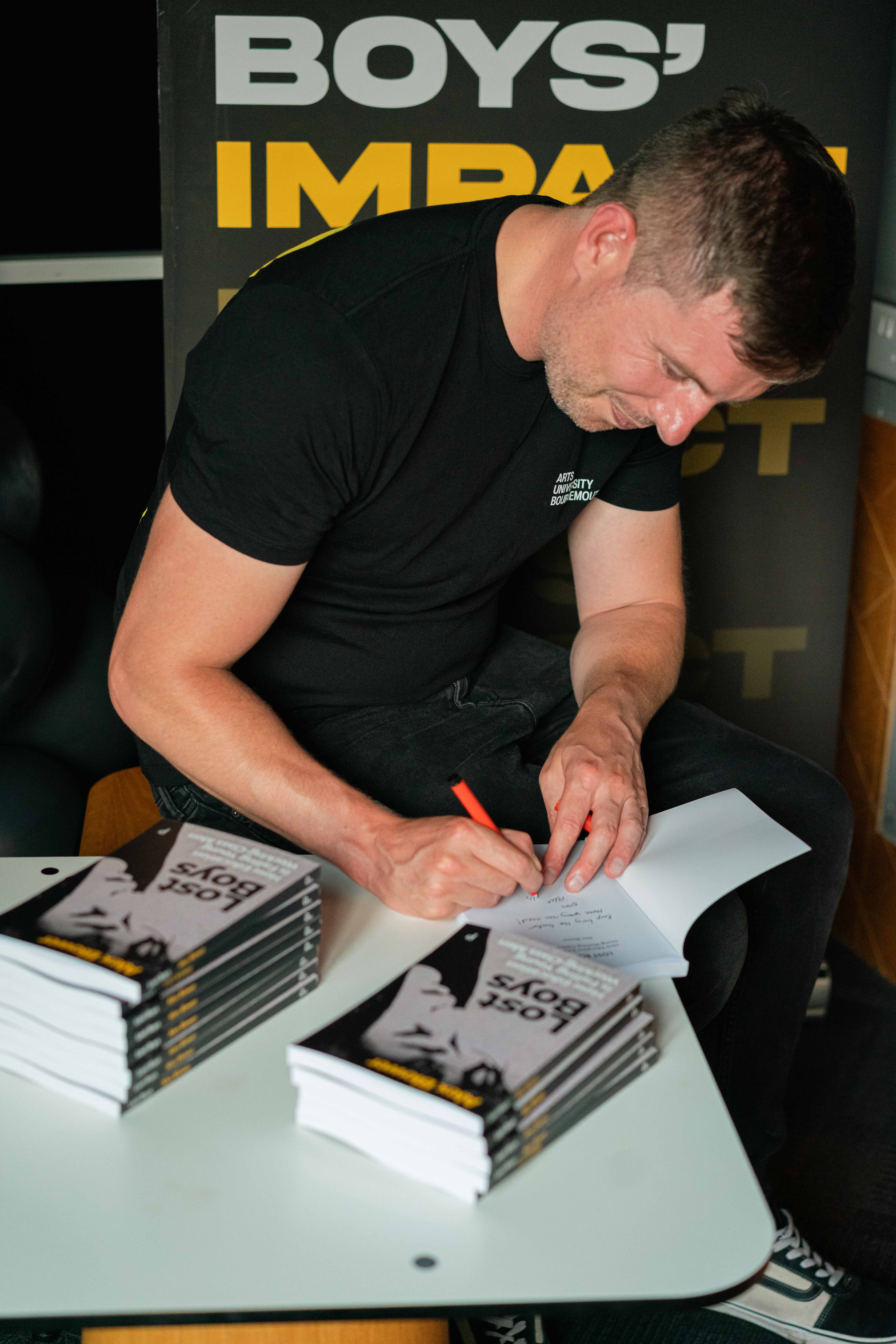 Dr A. Blower sitting and signing copies of his book "Lost Boys: How Education is Failing Young Working-Class Men". Two stacks of copies sit on a table in front of him.