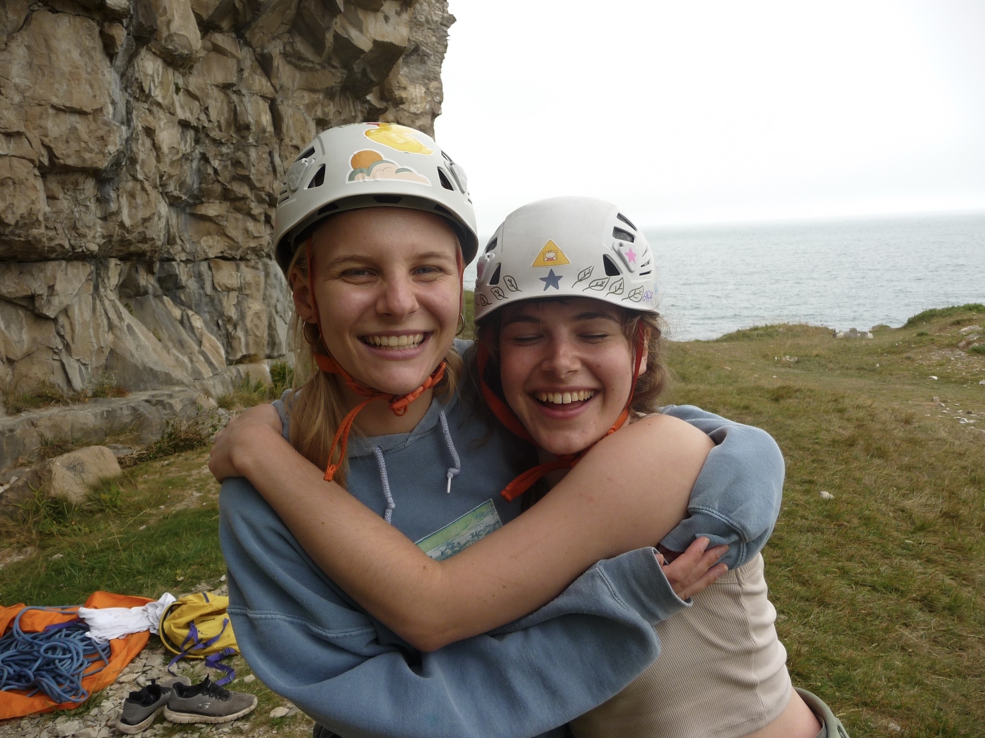 Two smiling people in white climbing helmets embracing near limestone cliff face with green countryside and sea visible behind them.