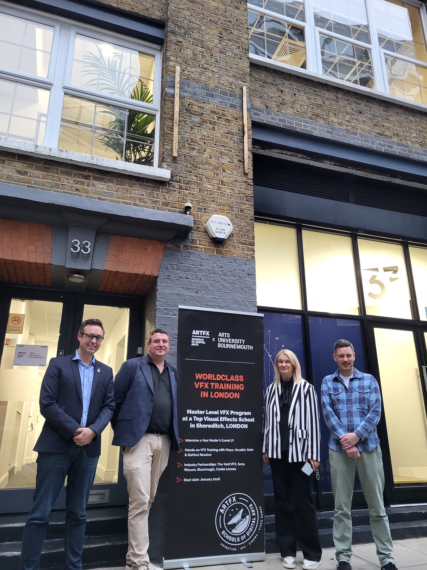 C Chorein, S Vanesse, L Mann, and J Carr standing beside black promotional banner outside brick building with large windows and number 33 entrance.