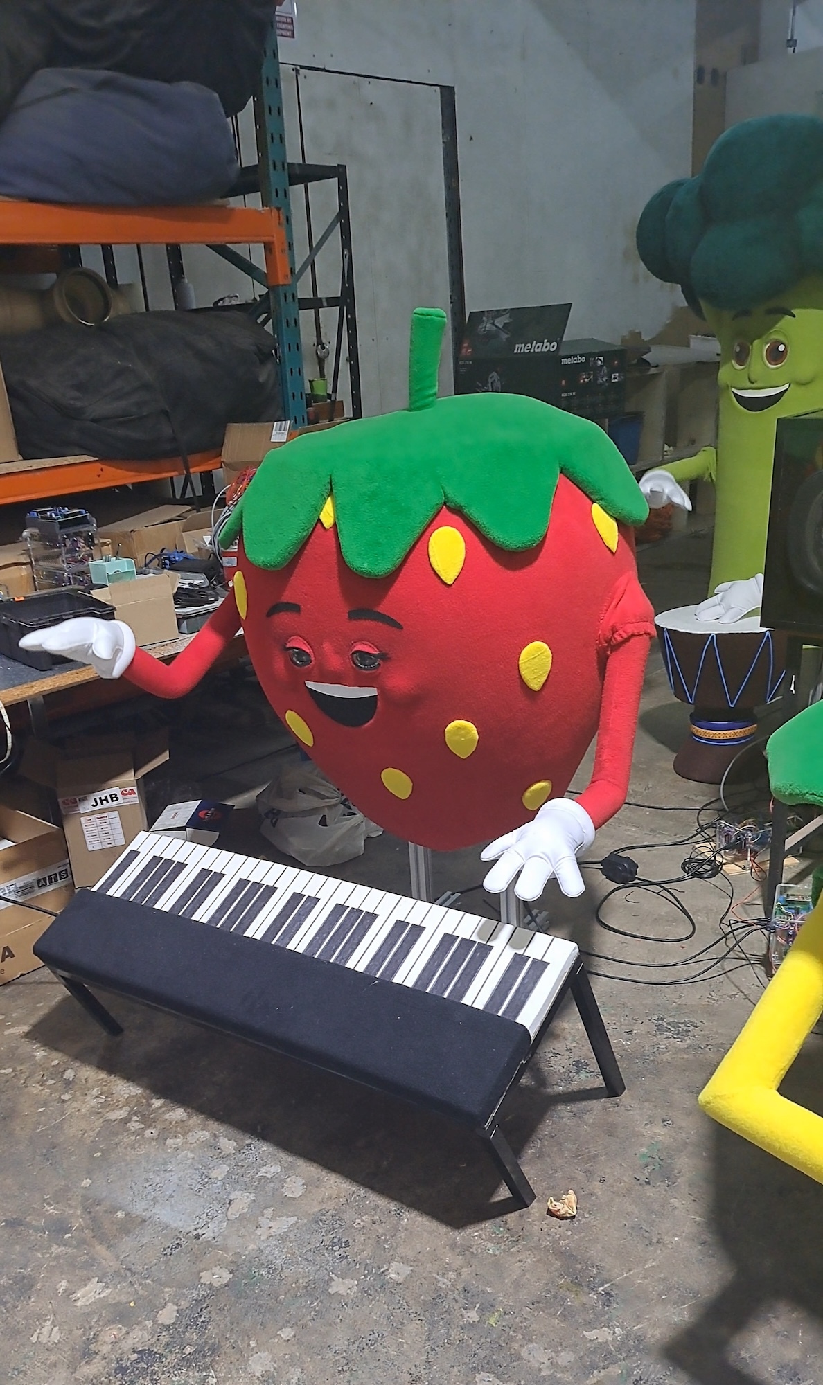 Red strawberry mascot with green leafy top, white gloves, standing behind black and white piano keyboard in cluttered workshop.