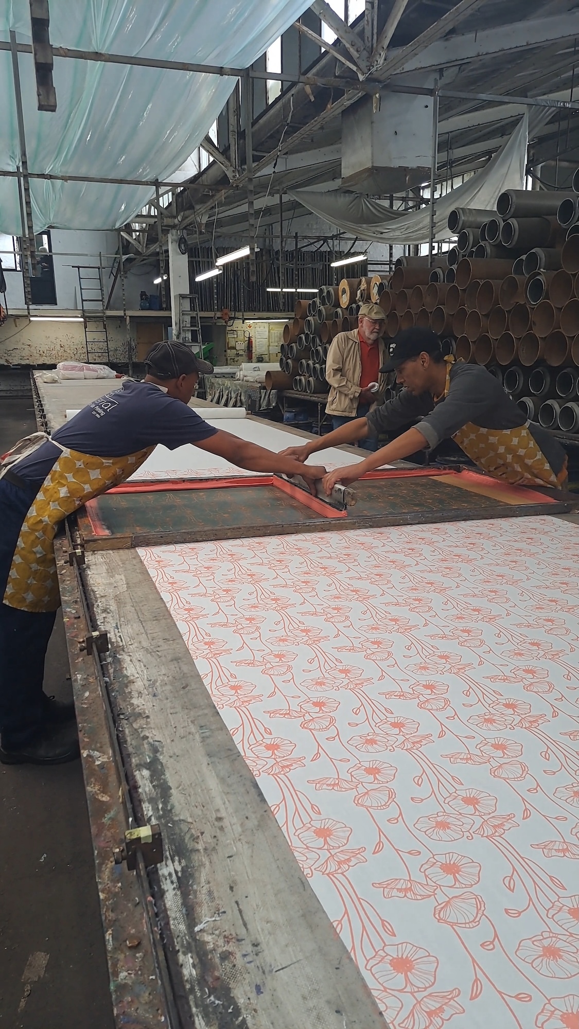 Two people screen-printing floral patterns onto fabric in industrial textile workshop with exposed metal beams and stacked tubes.