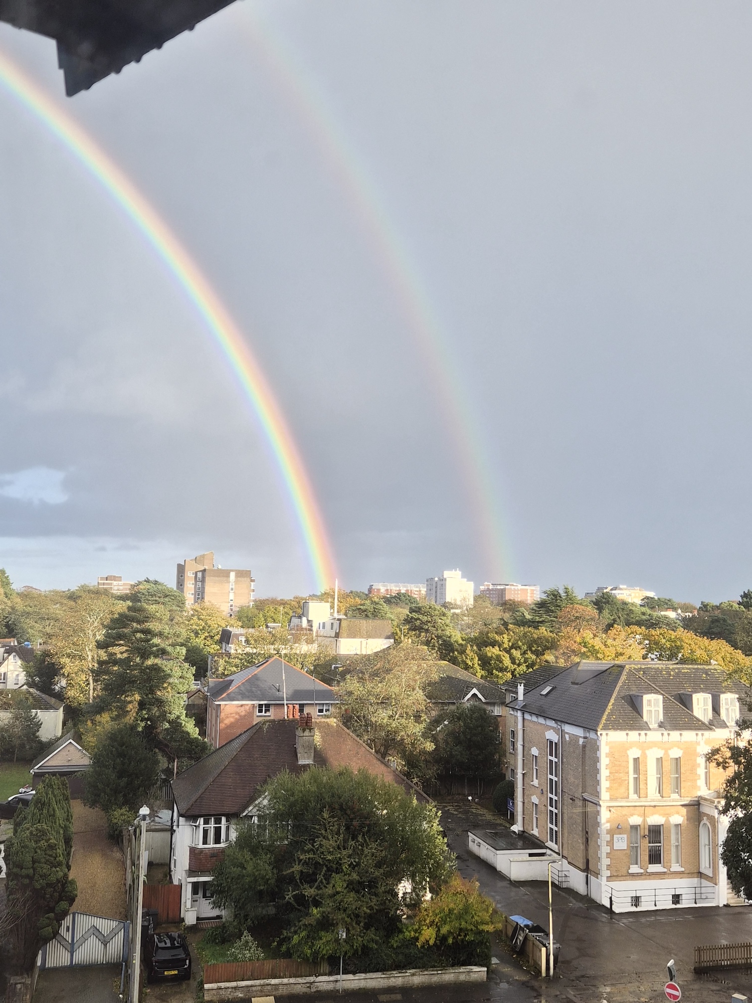 A vibrant double rainbow arcs across a cloudy sky over a residential area. Buildings and trees are visible in the foreground, with patches of green foliage and urban structures in view beneath the rainbows.