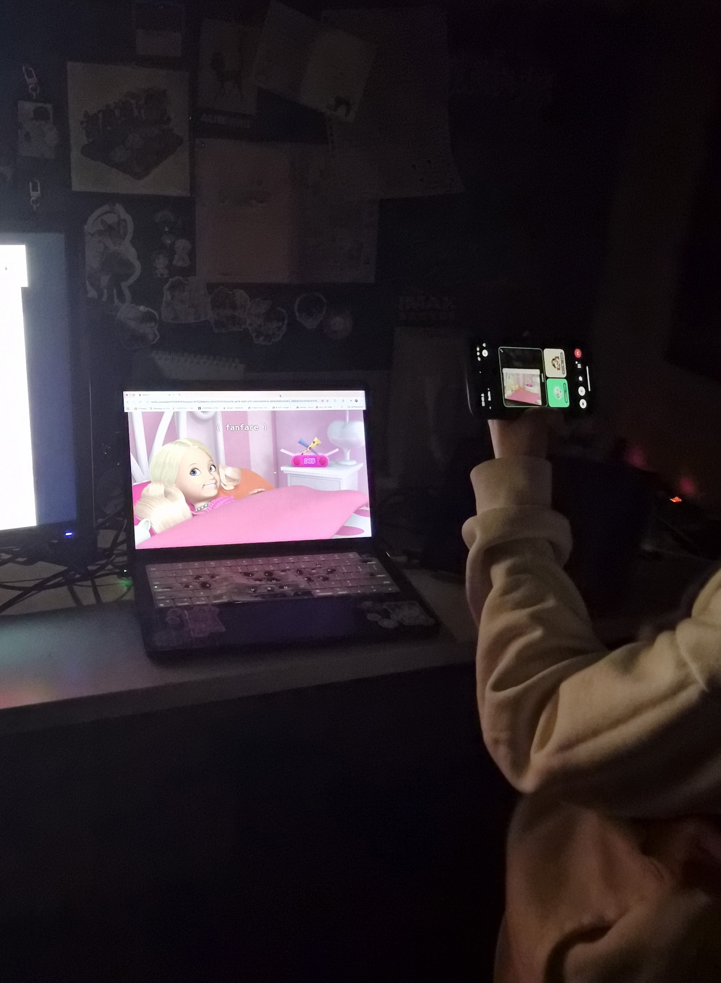 A person in a white sweatshirt holds a camera towards a laptop screen displaying a cartoon character in a pink room. The surrounding environment is dimly lit with a second computer monitor visible in the background.