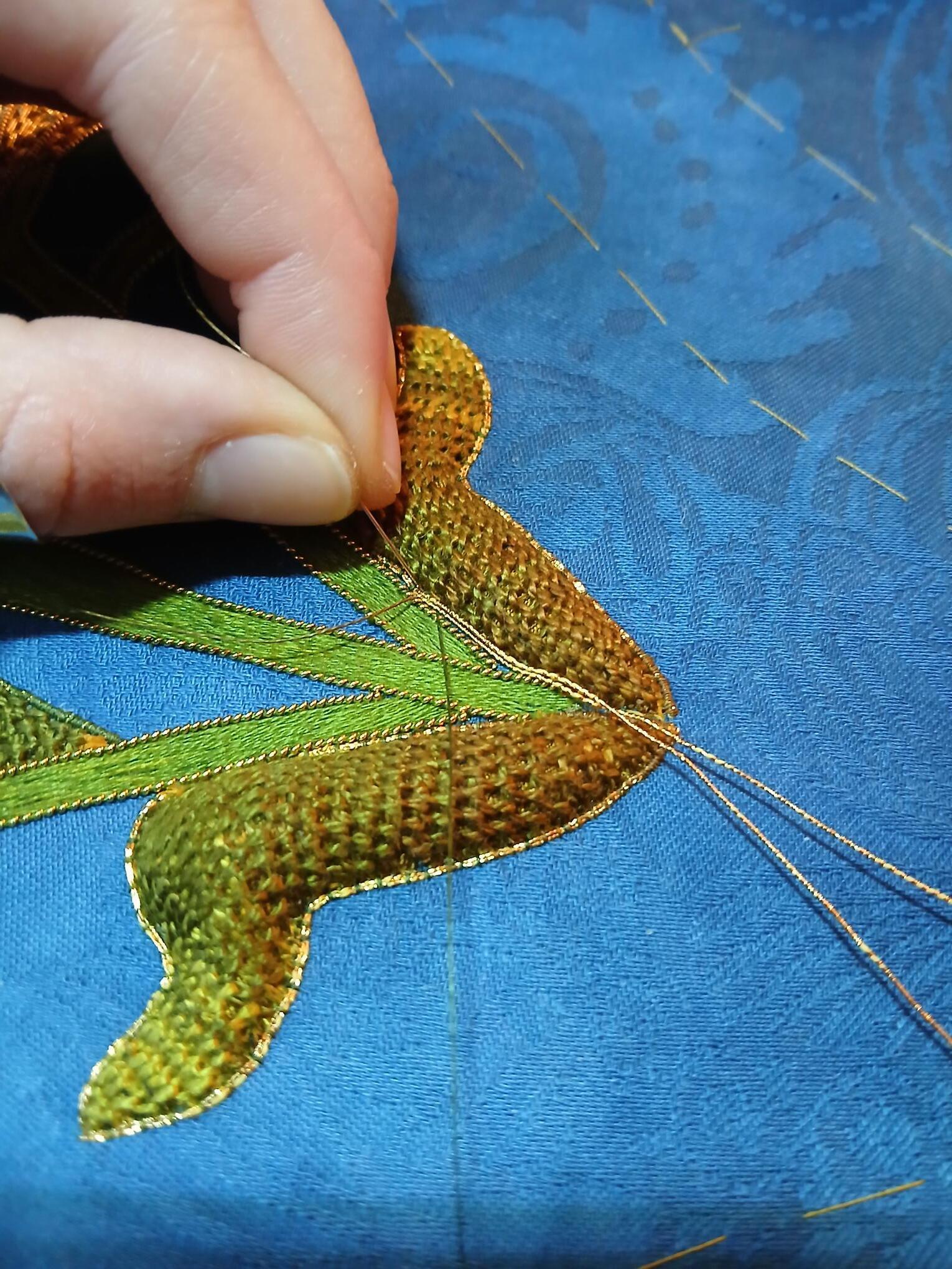 Fingers holding golden sequinned fabric flower with green leaves against blue silk material with diagonal stitching lines.