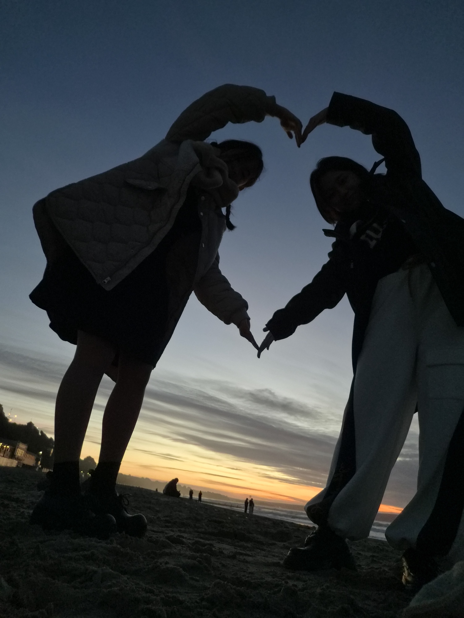 Two young people, silhouetted against a sunset sky, stand on a sandy beach and form a heart shape with their arms. The horizon displays vibrant colours, and distant figures can be seen walking along the shore.