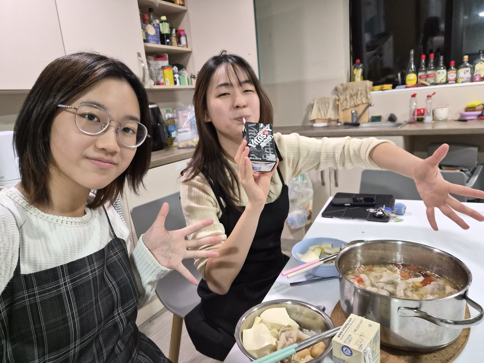 Two young people sitting at a table in a kitchen. One young person, wearing black glasses and a plaid dress, raises their hands, while the other holds a drink. A pot with stewing food and a plate of ingredients are on the table, with a background of kitchen shelves.