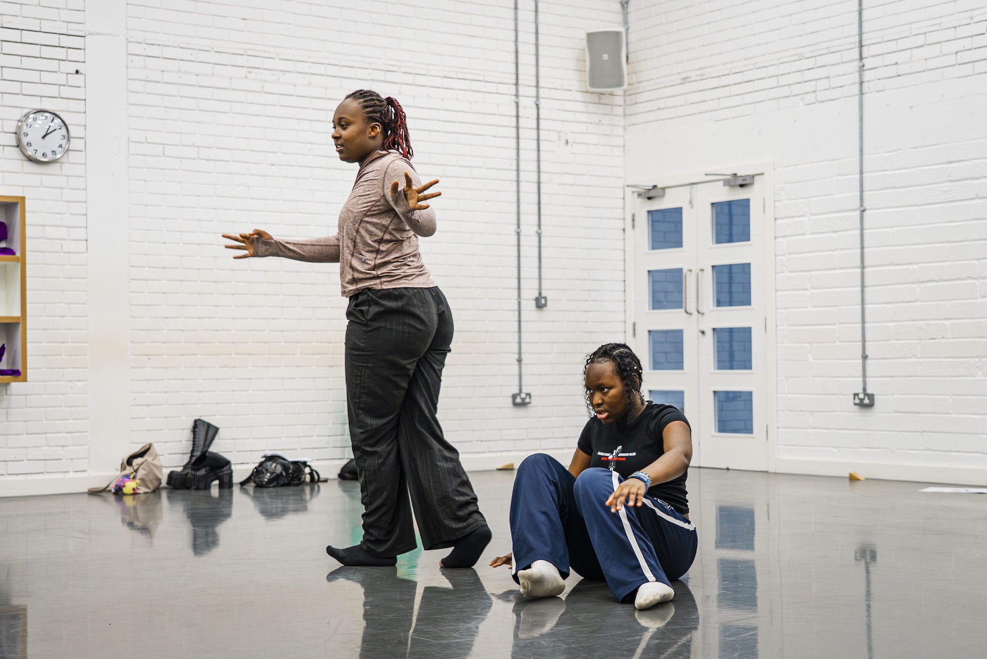 Two young people rehearse in a white studio with a polished floor, one standing with her arms extended and the other seated on the ground.
