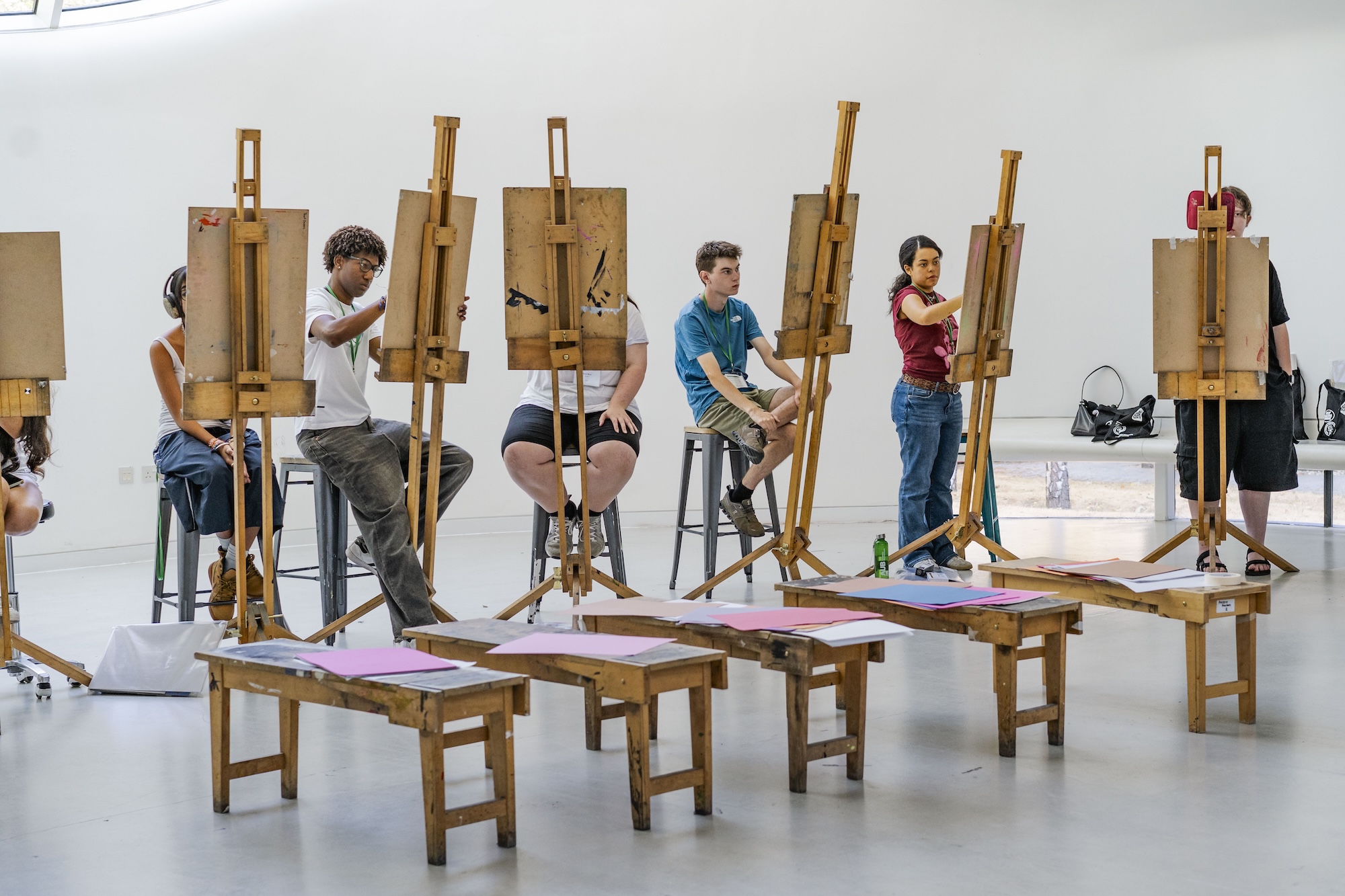 Several young people sit on stools and work at easels with wooden drawing boards in a bright studio, with low benches and art materials in the foreground.
