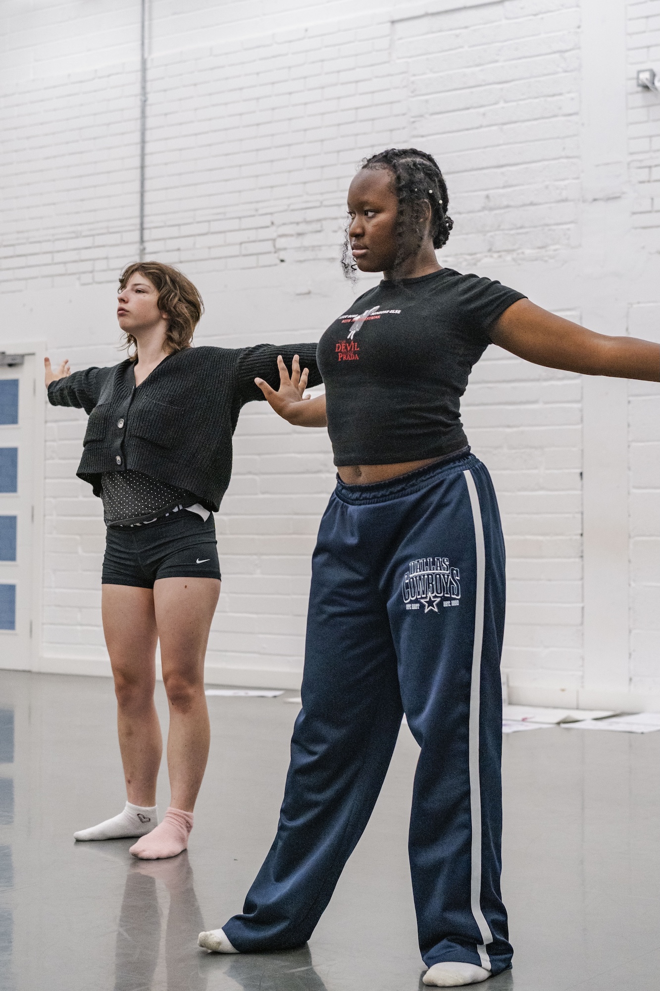 Two young people stand with arms outstretched in a dance studio with a white brick wall and reflective floor.