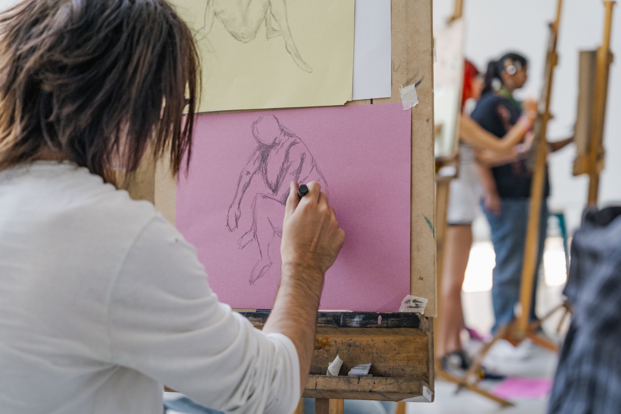 A person sketching on pink paper at an easel, with another artist painting in the background.