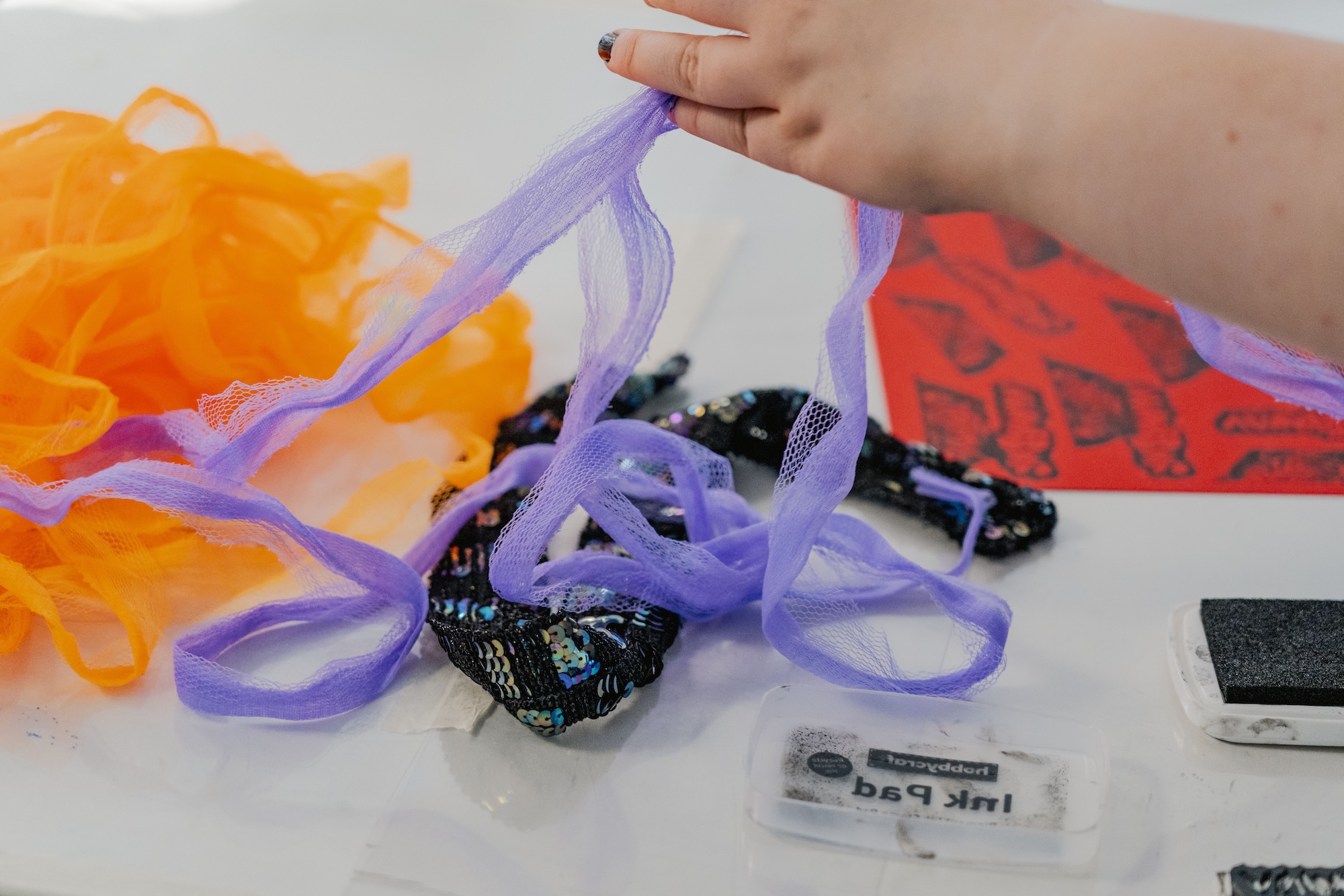 A person's hand holds a purple mesh fabric strip above a table with orange netting, a sequinned pouch, and a red paper sheet.