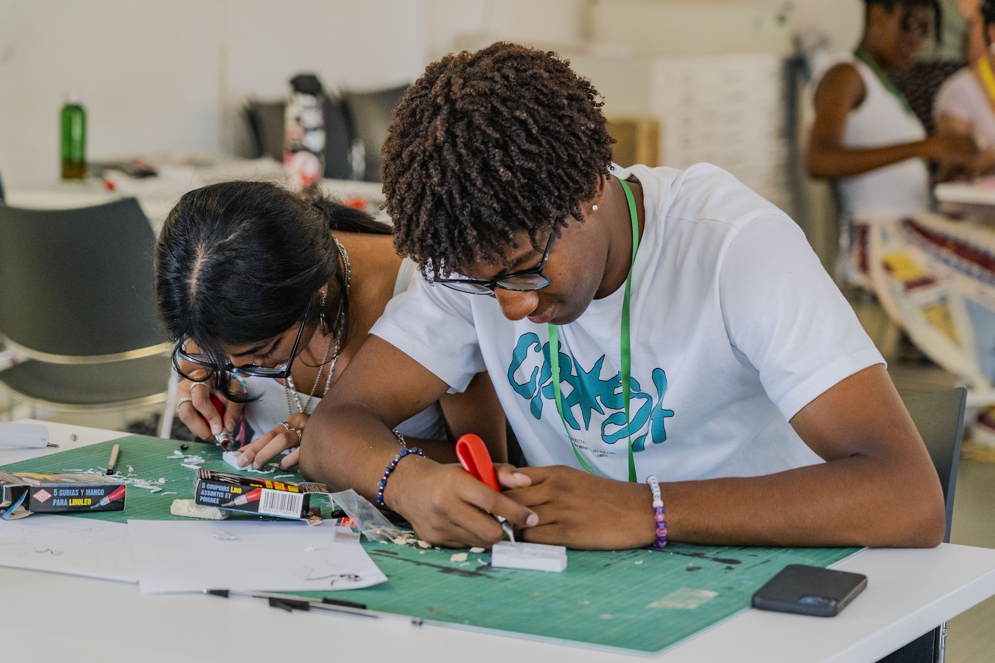 Two young people work on a craft project at a table covered with tools, paper and a green cutting mat.