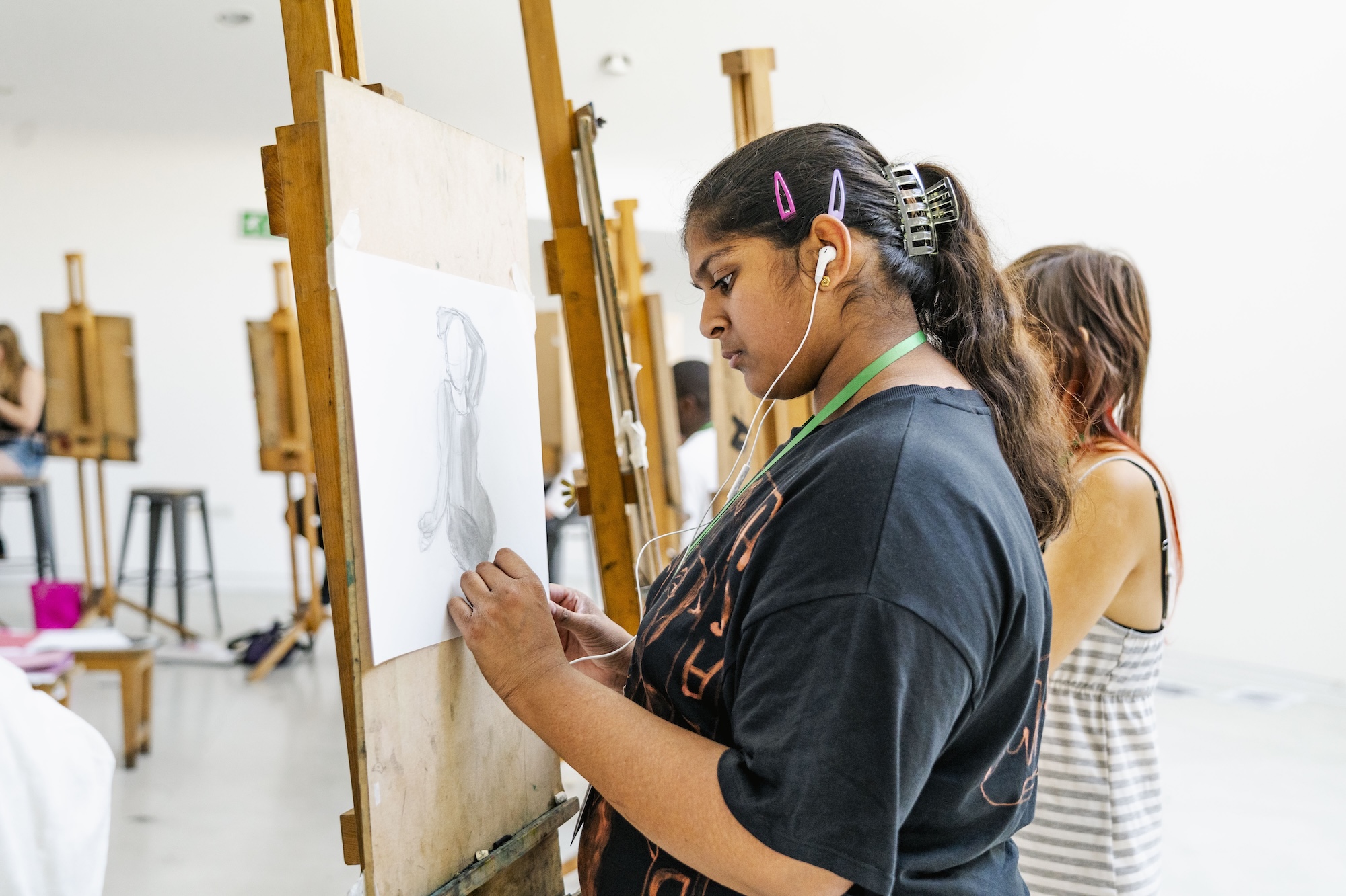 A young person sketches on a sheet of paper clipped to a wooden easel in an art studio, with other easels and students visible in the background.